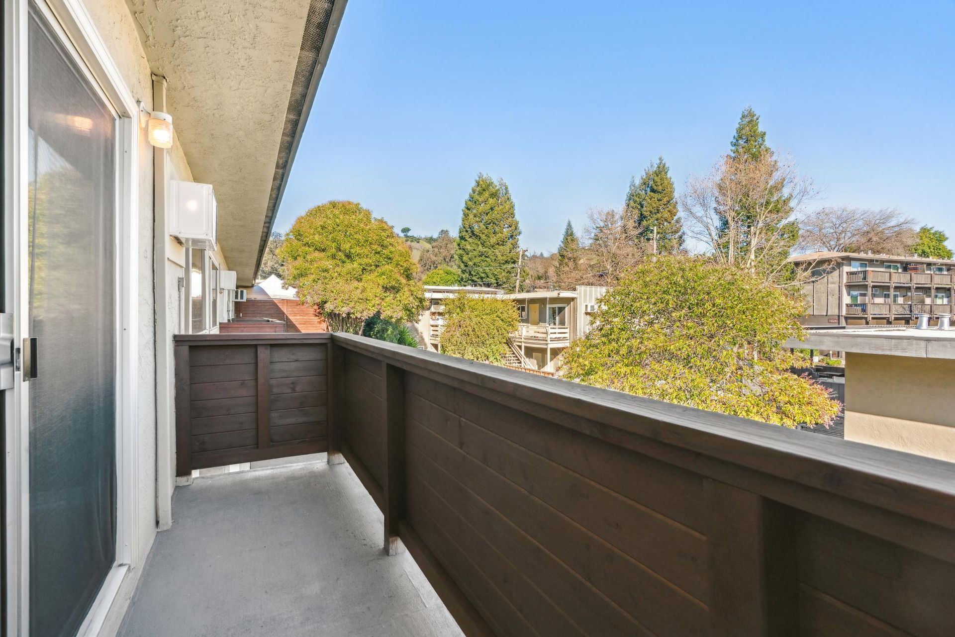 Balcony with wooden railing overlooking trees and buildings on a sunny day.