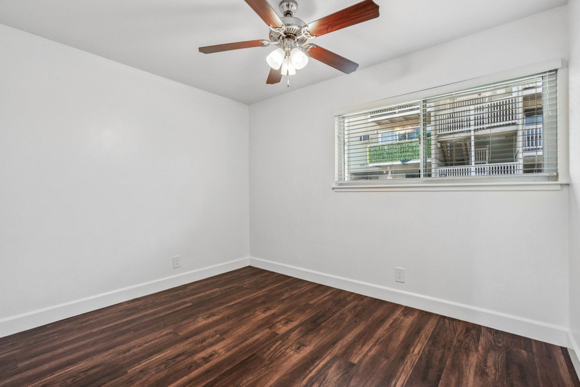 Empty bedroom with wood-look floor, white walls, ceiling fan, and window with blinds.