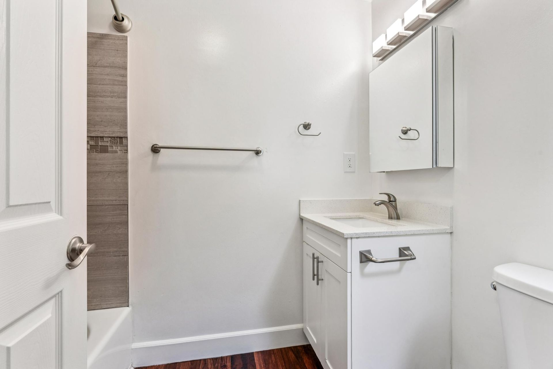 White bathroom with shower, vanity, and toilet; dark wood floor.