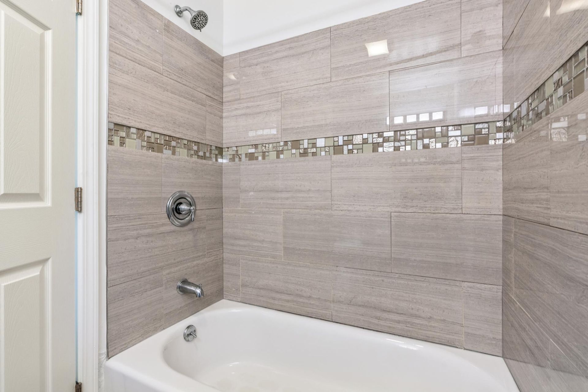 Bathroom with gray tiled shower wall and white bathtub. Silver fixtures and a decorative mosaic band.
