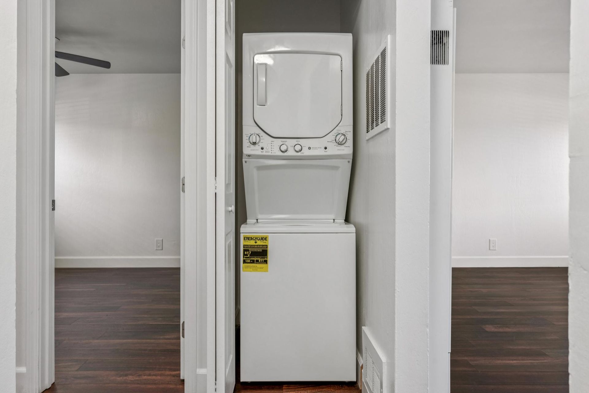 Stacked white washer and dryer in a hallway between two rooms with hardwood floors.