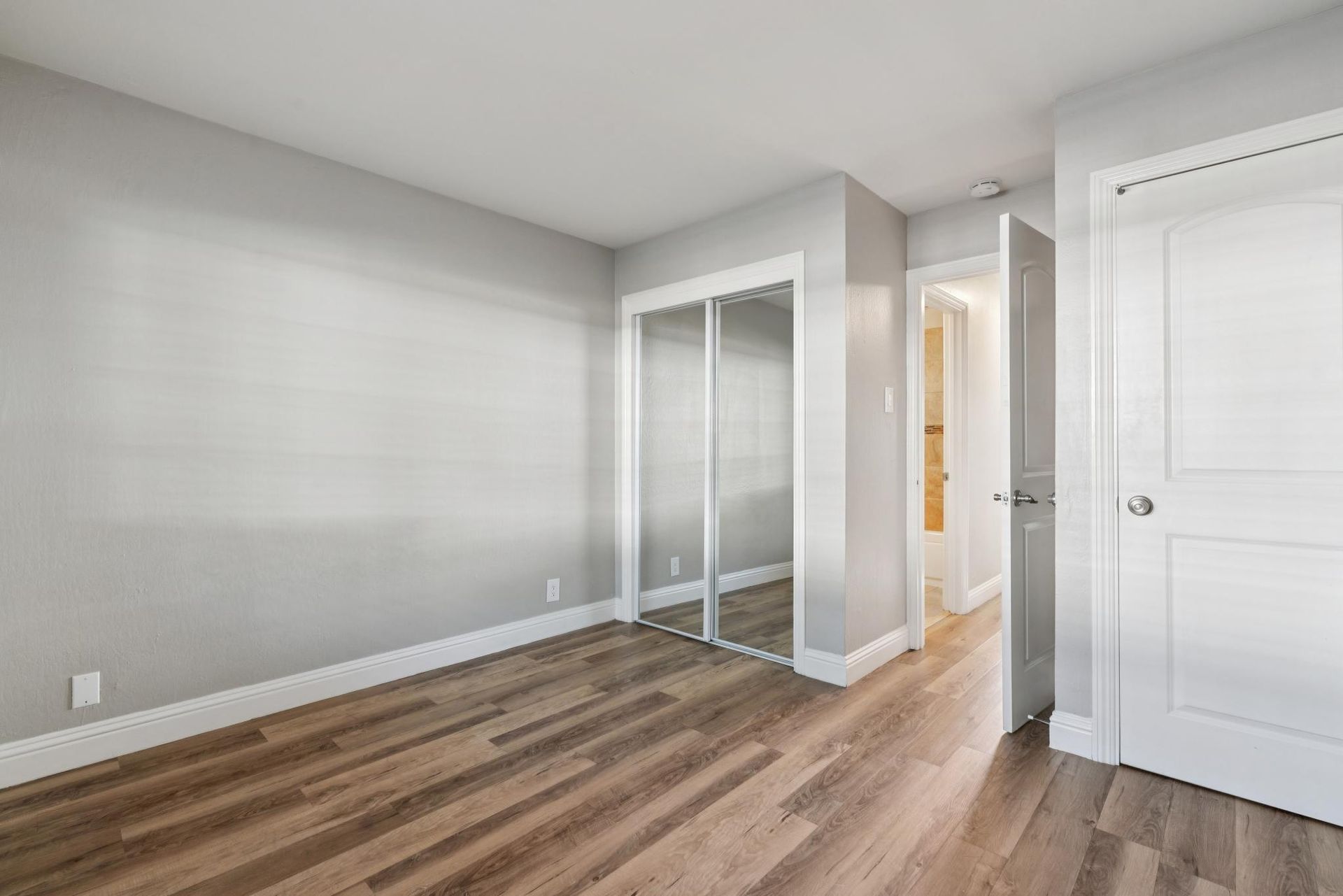 Empty bedroom with hardwood floors, two mirrored closet doors, and a hallway entrance.