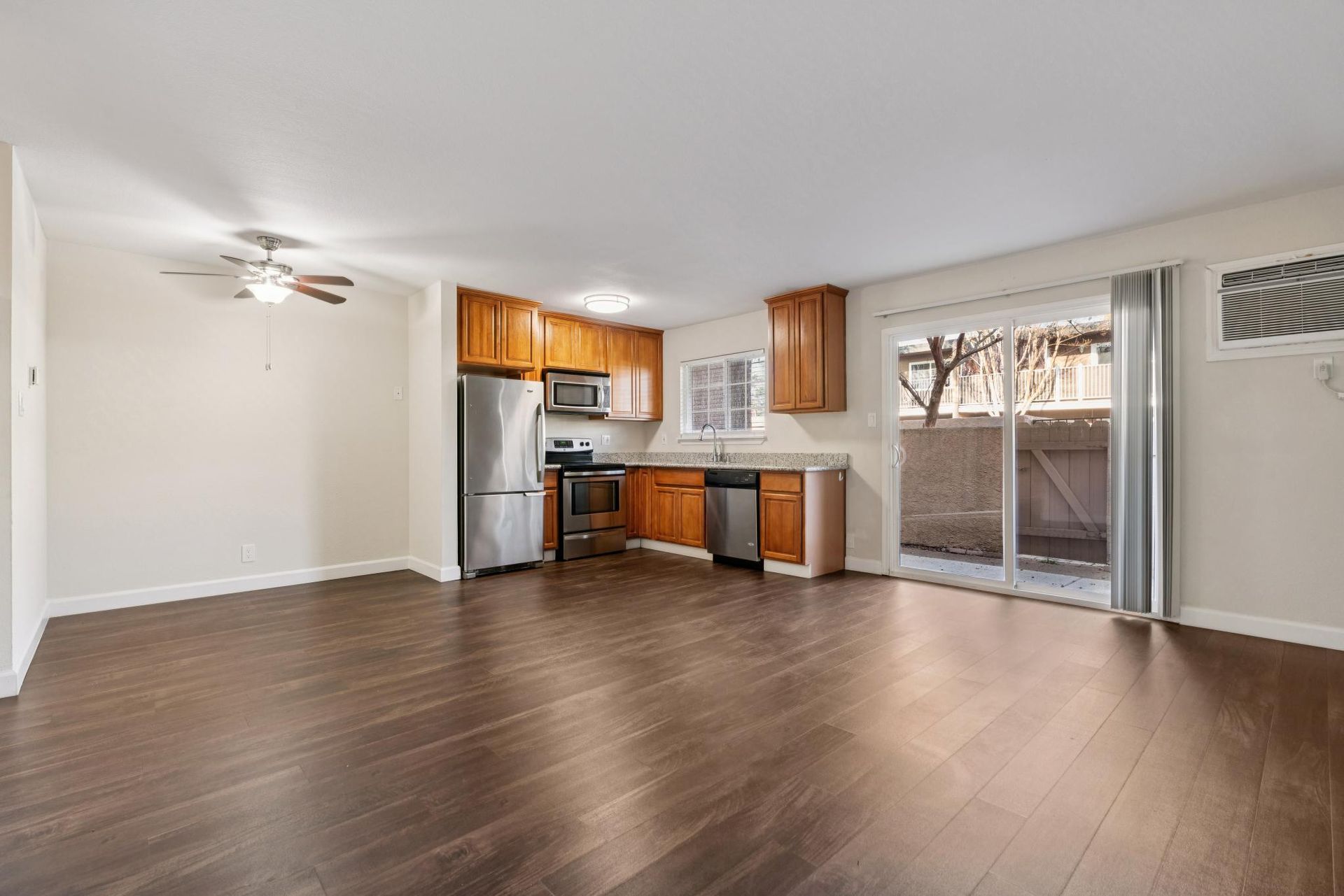 Empty living room and kitchen with hardwood floors, stainless steel appliances, and sliding glass door to a patio.