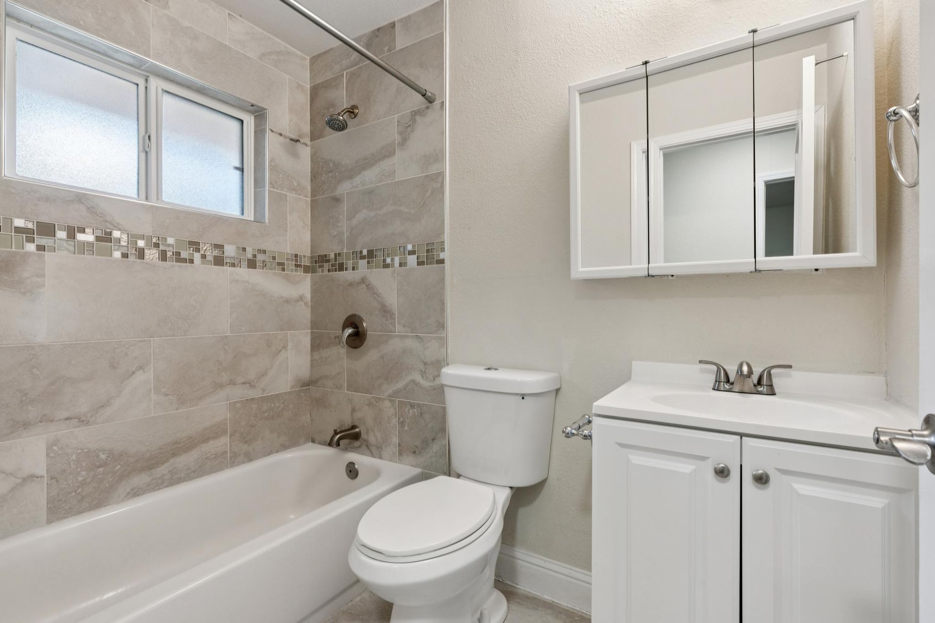 Bathroom with tub, toilet, and vanity. Gray and white tiles, medicine cabinet, and window.