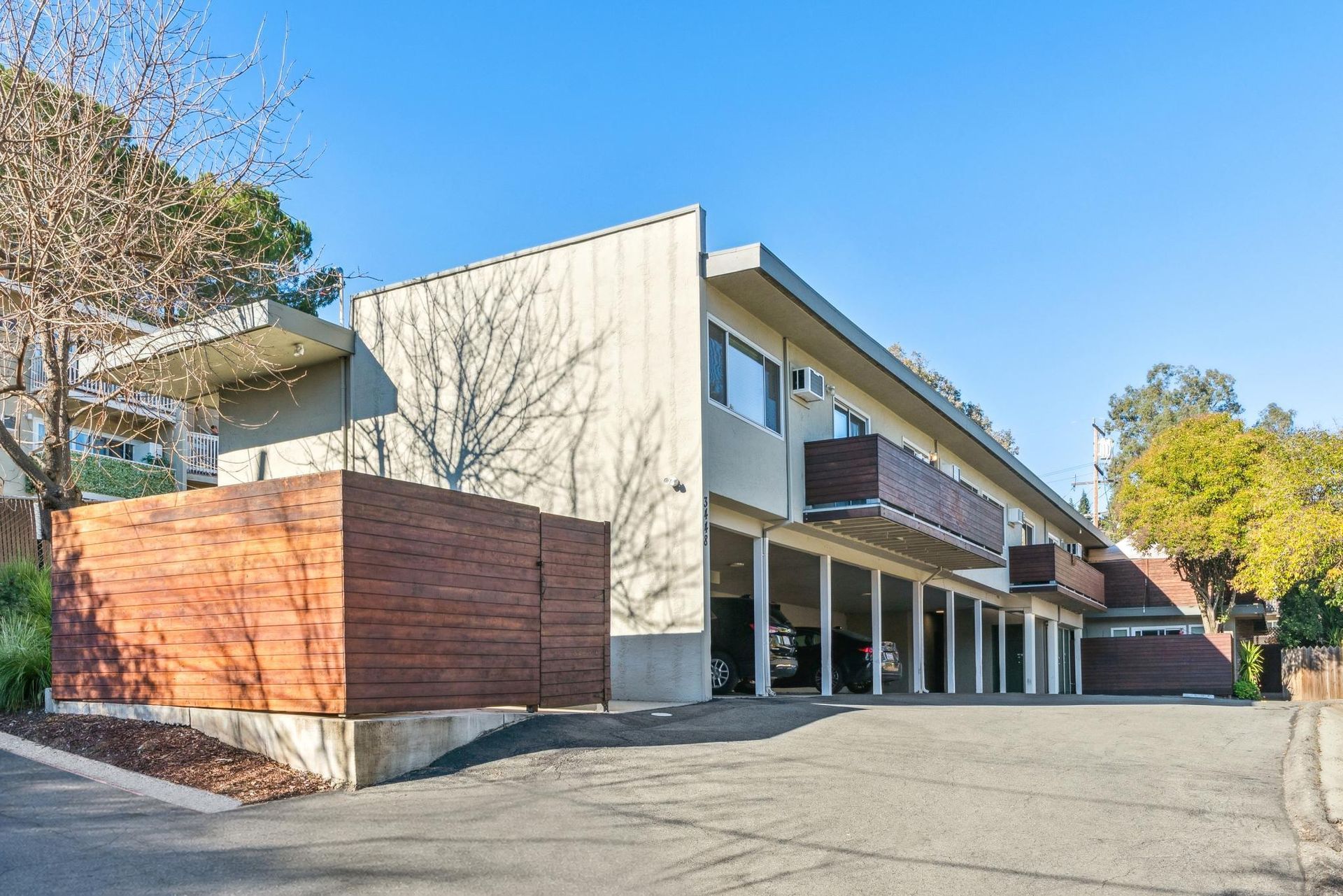 Two-story apartment building with covered parking, balconies, and a wooden privacy fence, under a blue sky.