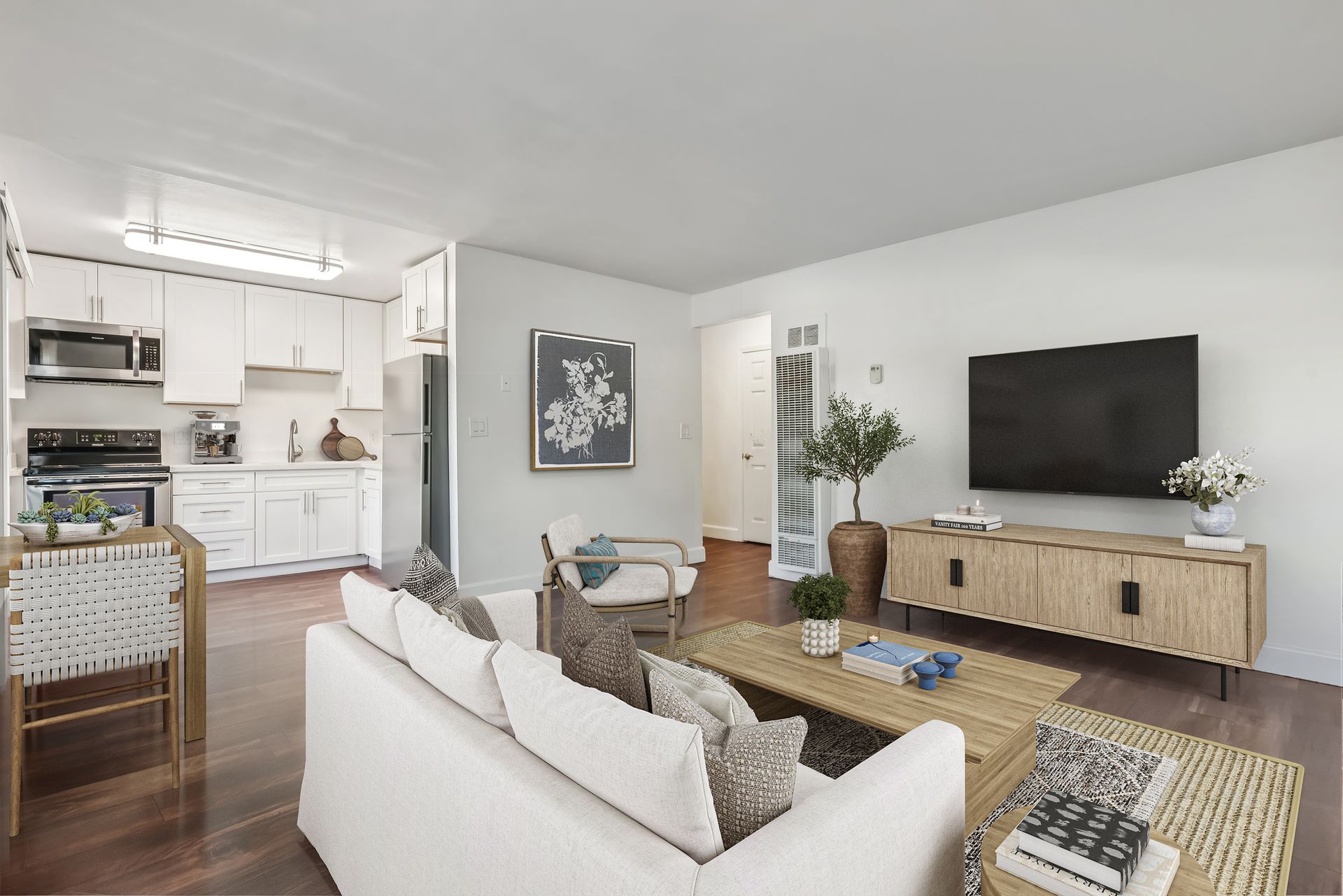 Living room with white sofa, wood flooring, and kitchen in the background. Television mounted on a light wood cabinet.