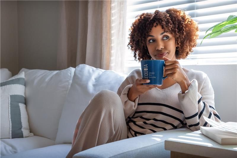 Woman sits on a white couch, sipping from a blue mug. She looks thoughtful, near a sunny window.