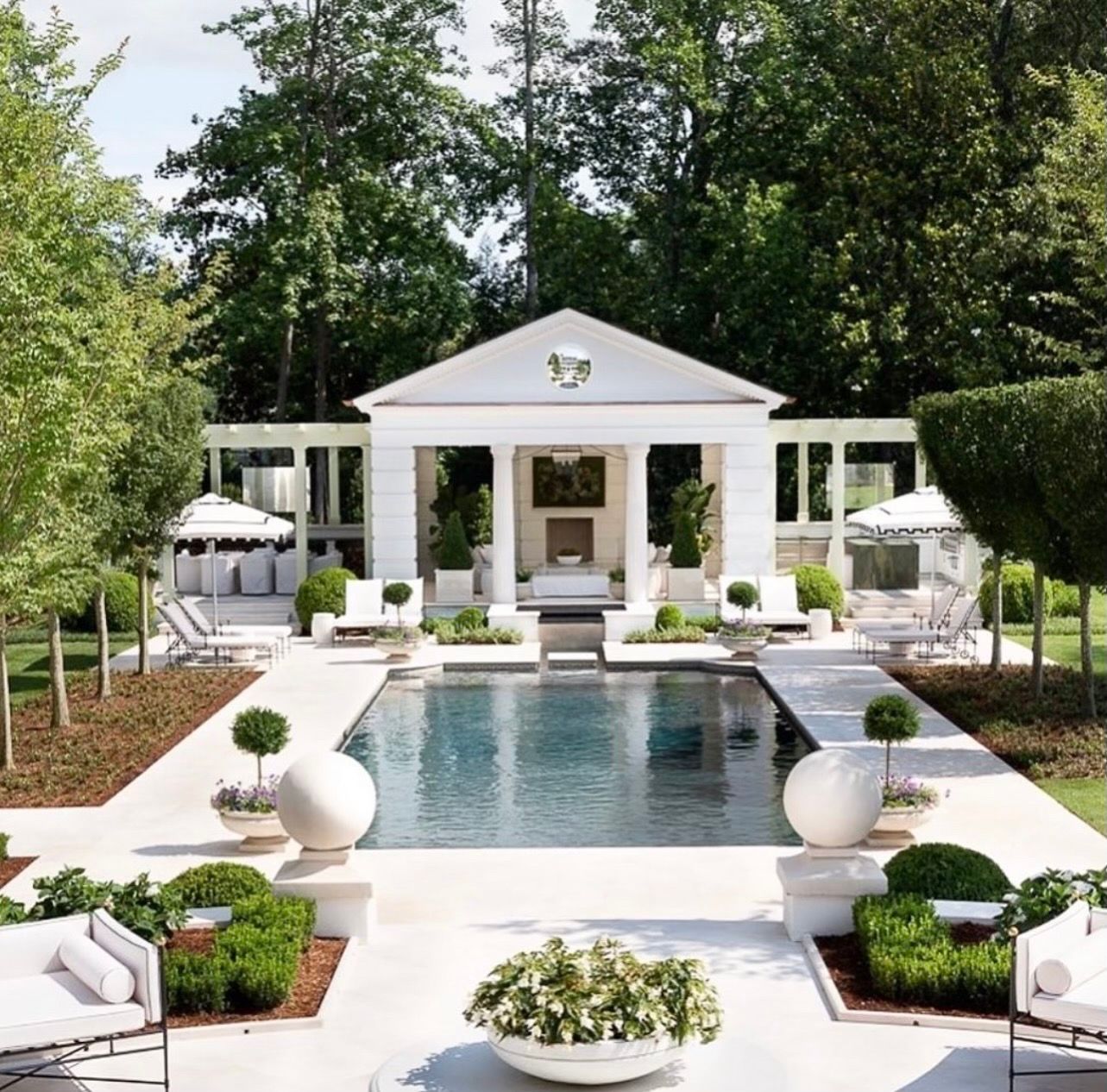 White pool house with columns and pool, flanked by seating areas and manicured landscaping.