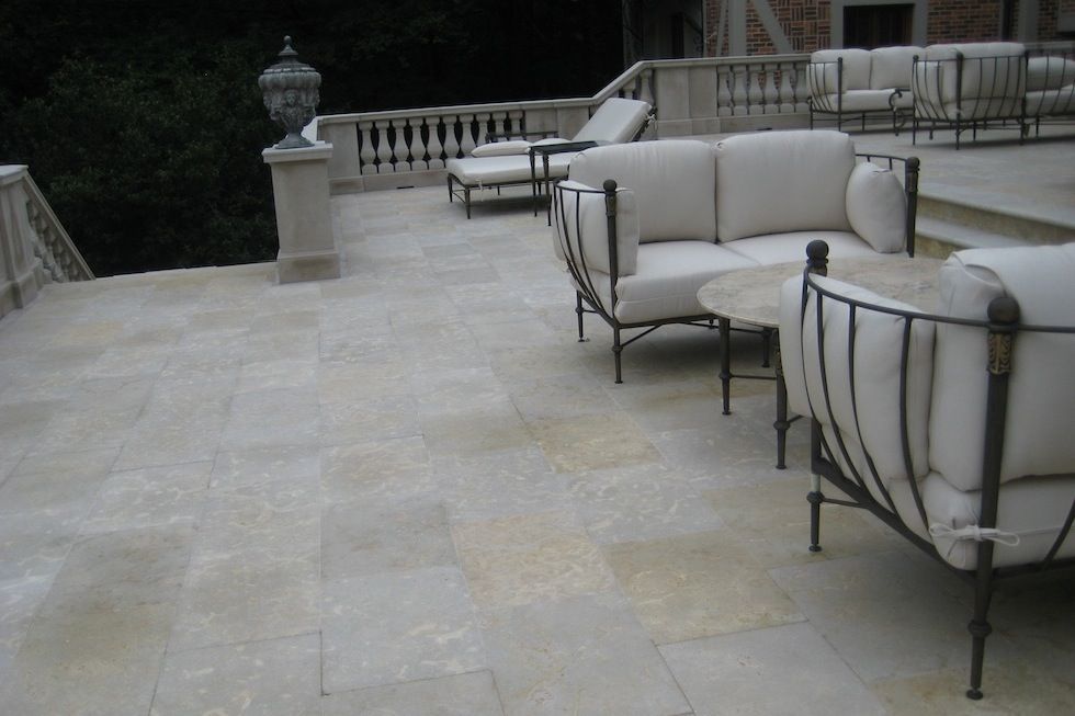 A stone patio with beige furniture: couches, chairs, and a table. In the background are railings and trees.