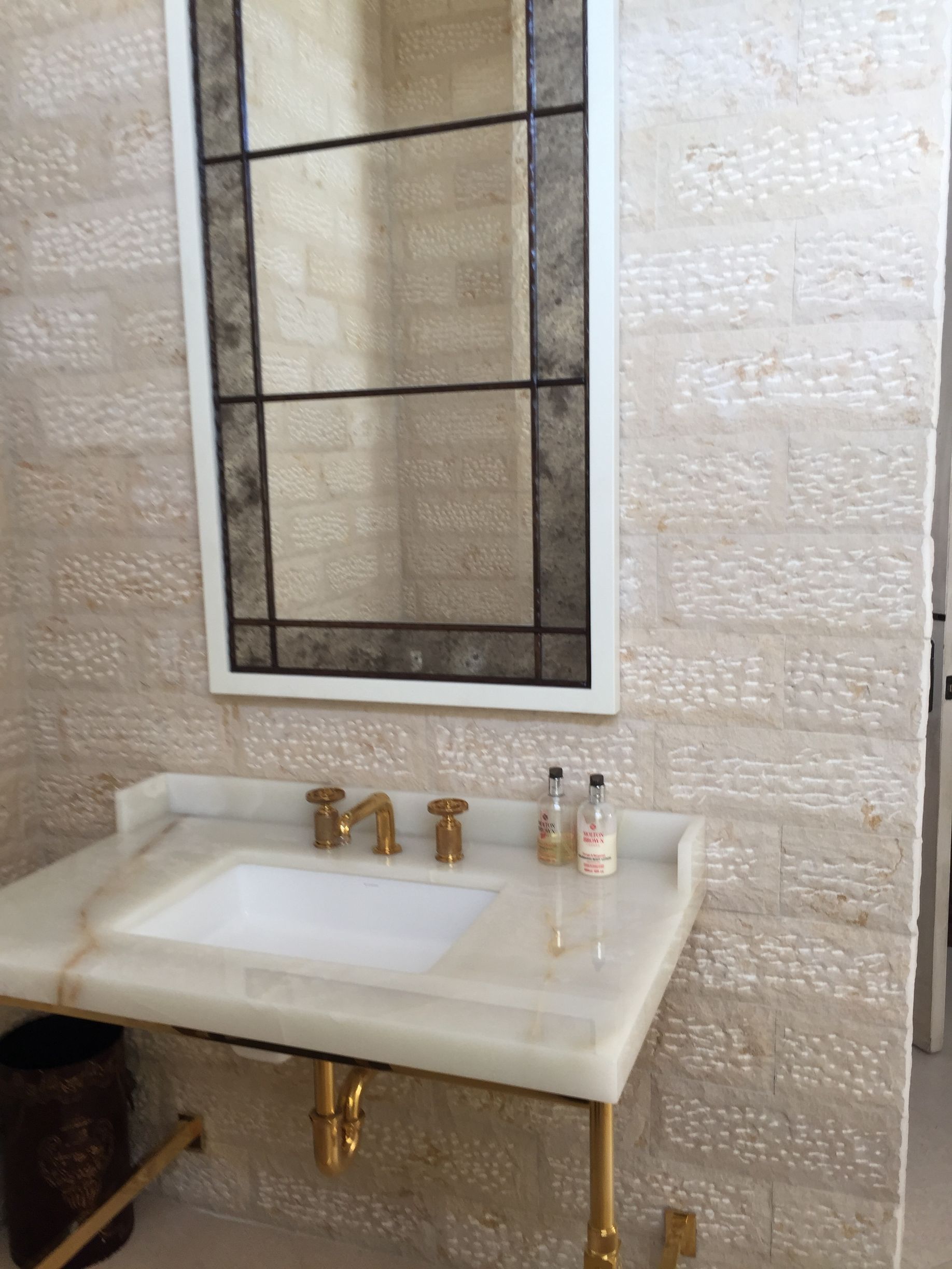 Bathroom with gold fixtures, white sink, mirror with dark trim, and textured beige walls.
