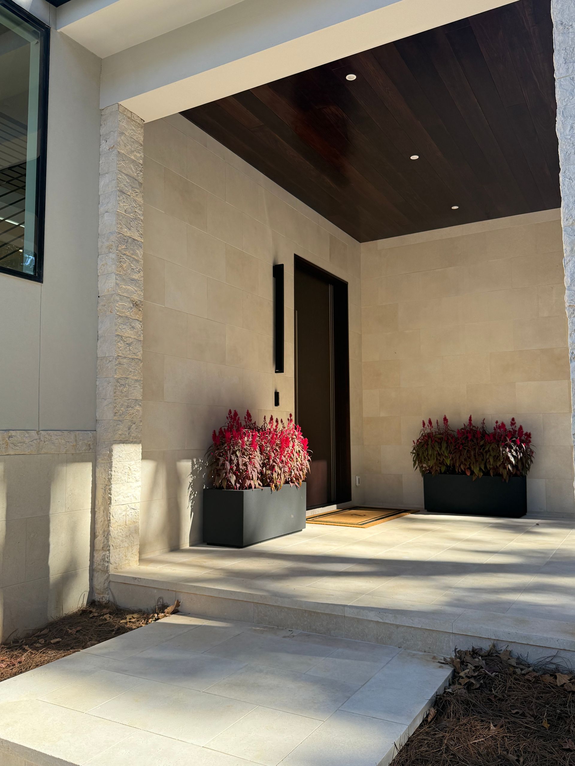 Covered porch with two planters of red flowers, dark wood ceiling, and a brown door.