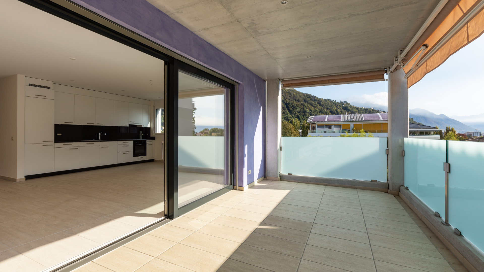 Balcony with tile floor, glass railing, and mountain view; open sliding glass door leads to modern kitchen.