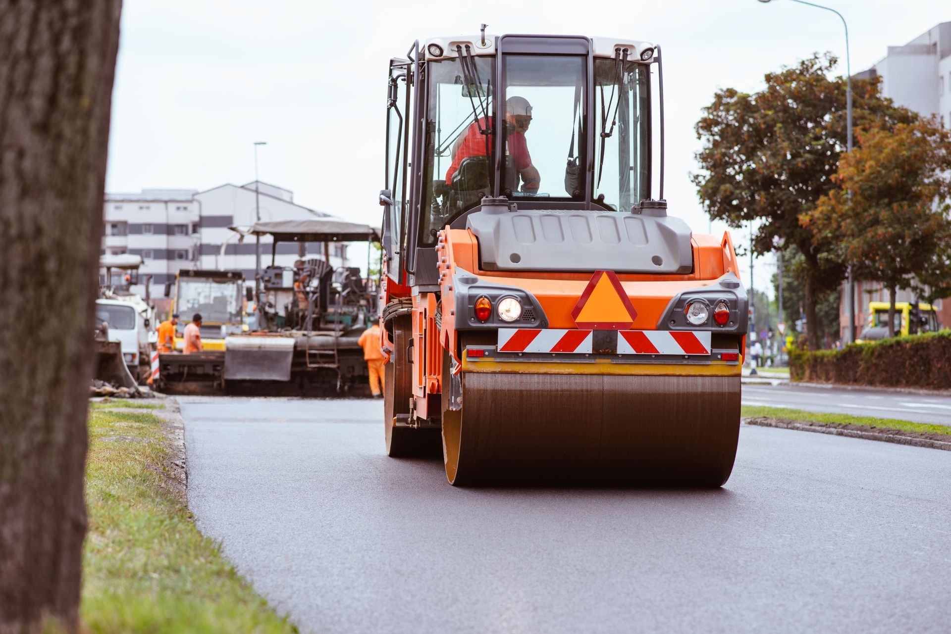 Road roller compacting freshly laid asphalt on a street, with construction workers nearby.