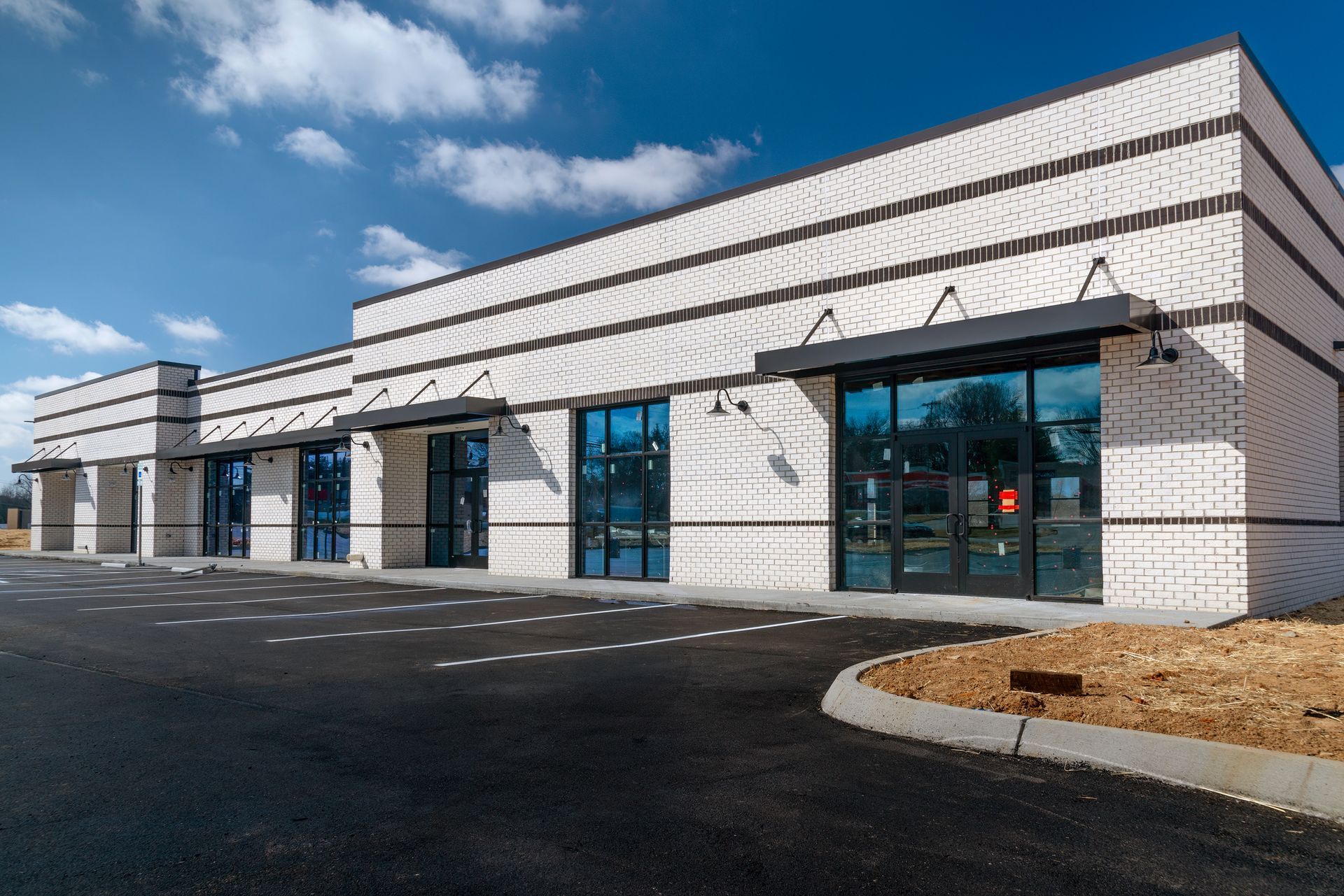 Modern brick building with large windows, awnings, and a black asphalt parking lot.