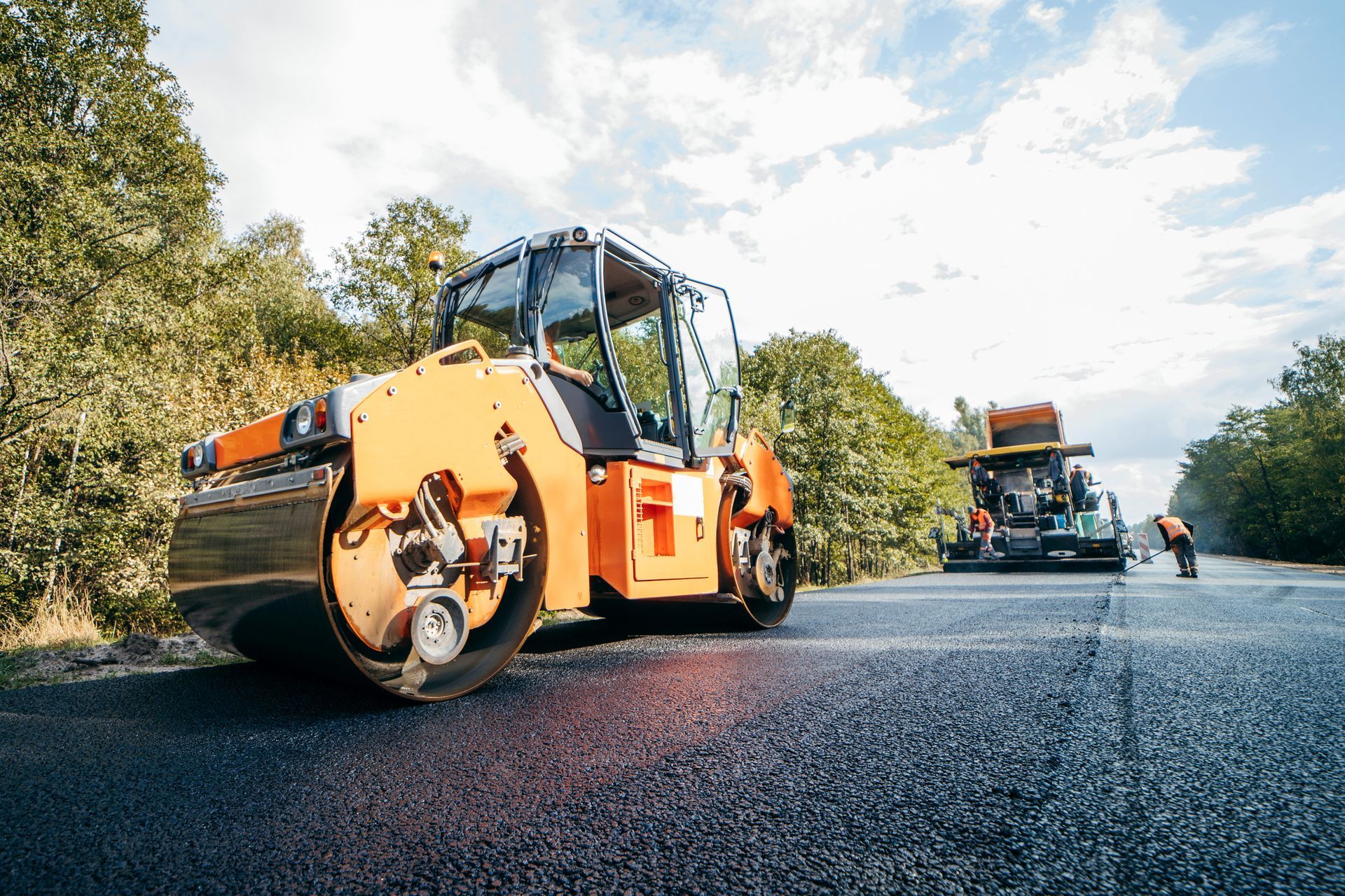 Orange road roller compacting fresh asphalt on a road, with trees in the background.