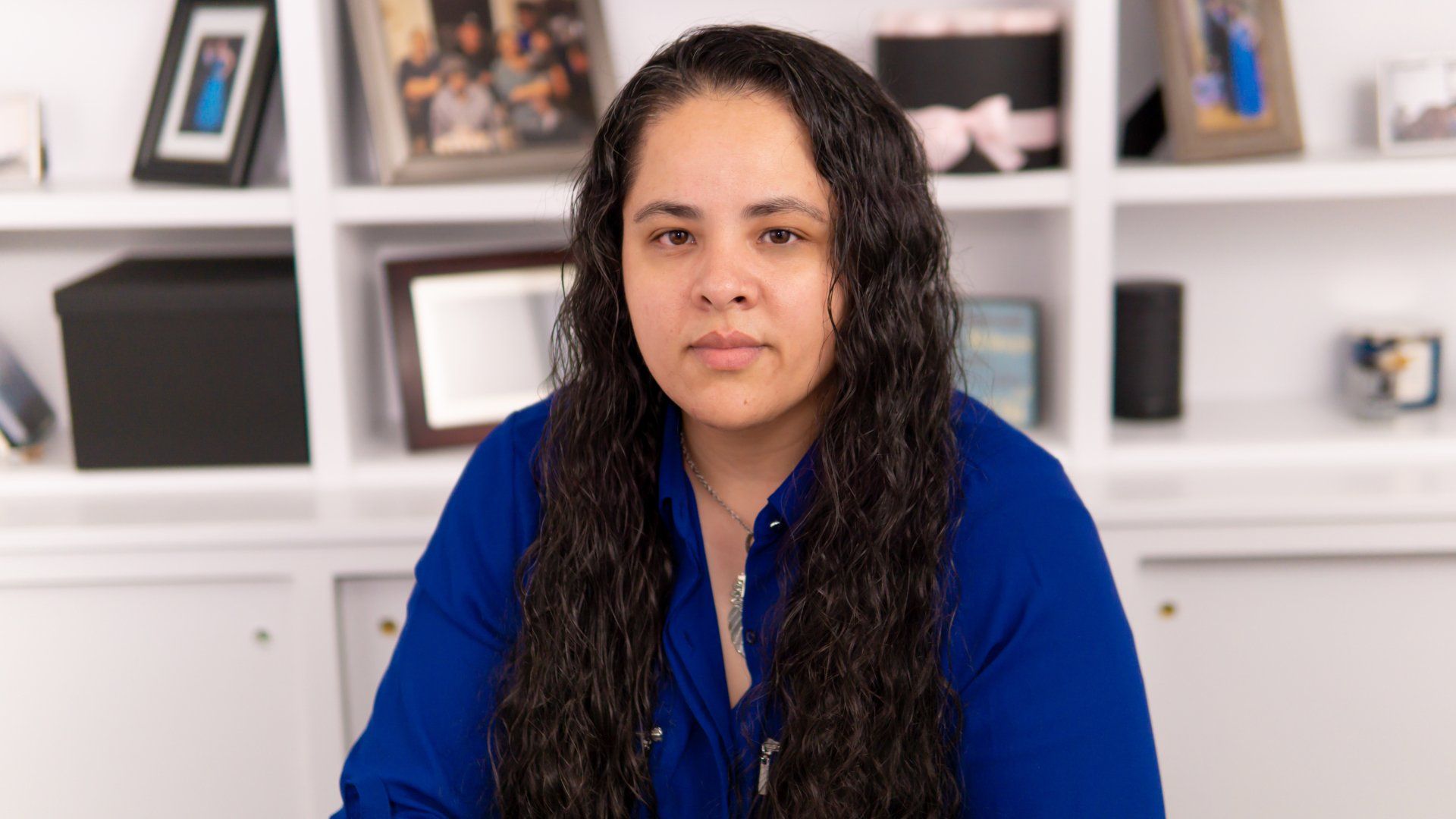 A woman in a blue shirt is sitting in front of a bookshelf with pictures on it.