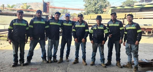Un grupo de ocho personas vestidas con uniformes de trabajo oscuros idénticos y franjas reflectantes de alta visibilidad, de pie en un patio al aire libre.