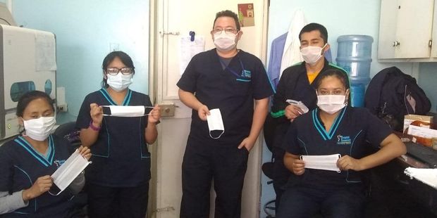 Cinco trabajadores sanitarios con uniformes azules y mascarillas posan en una oficina, sosteniendo mascarillas quirúrgicas.
