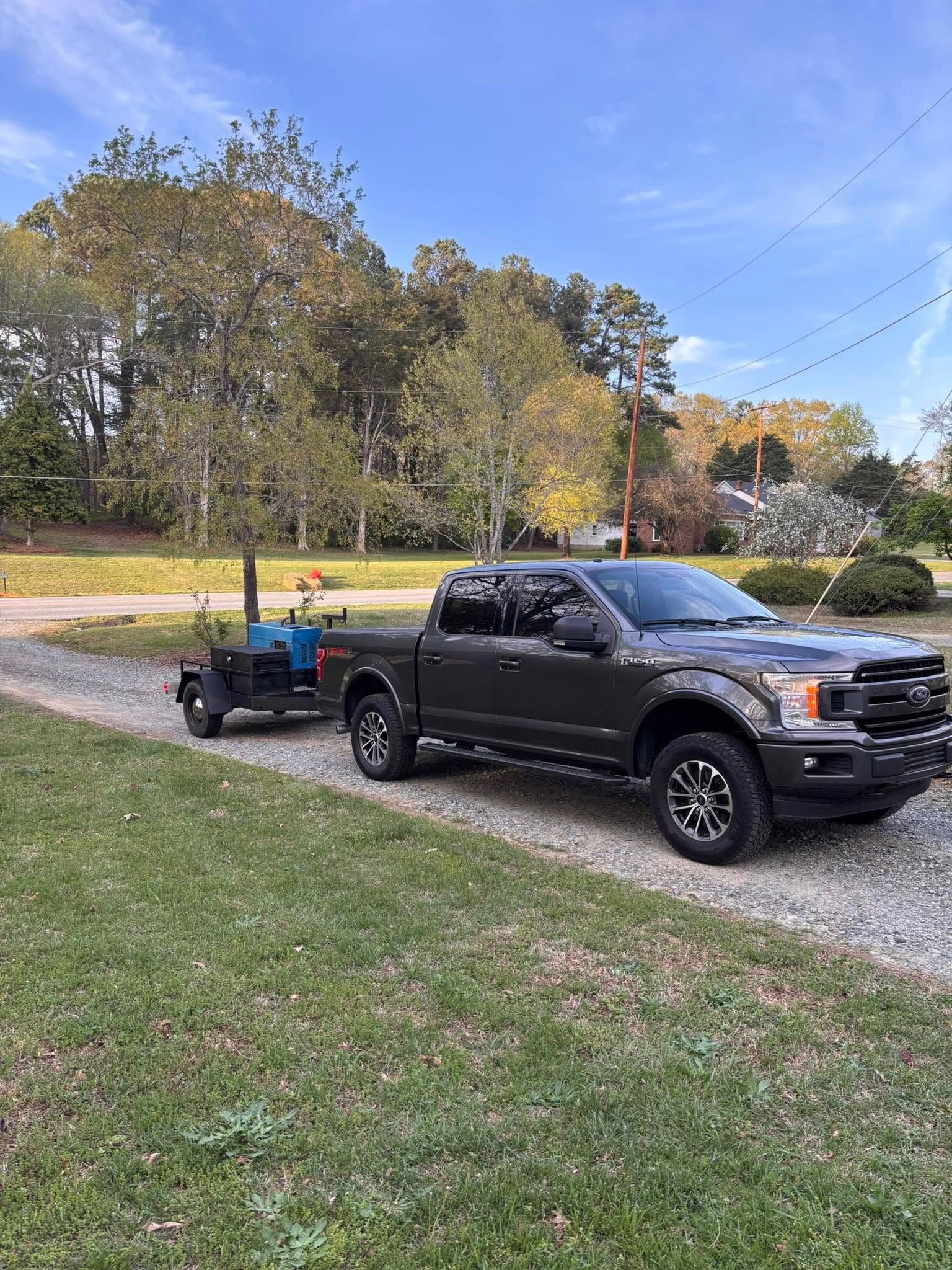 Dark gray pickup truck towing a blue smoker on a gravel driveway, trees in the background.