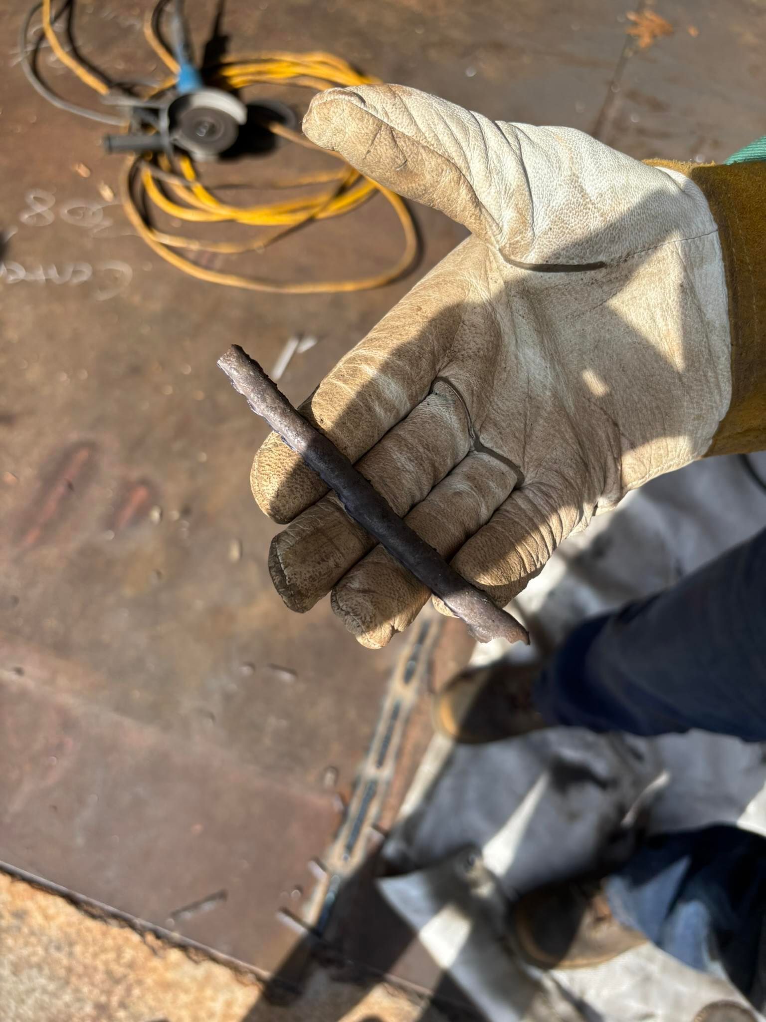 Gloved hand holding a dark metal rod, possibly rebar, with an angle grinder in the background.