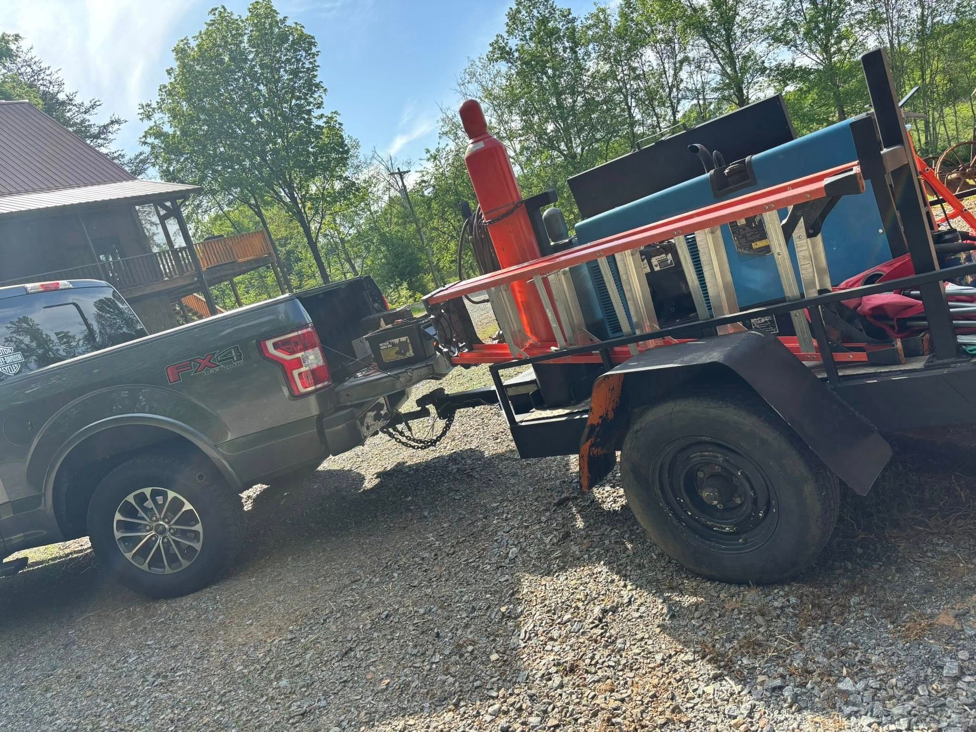 Gray pickup truck towing a trailer with a ladder, tank, and tools. Outdoors on gravel.