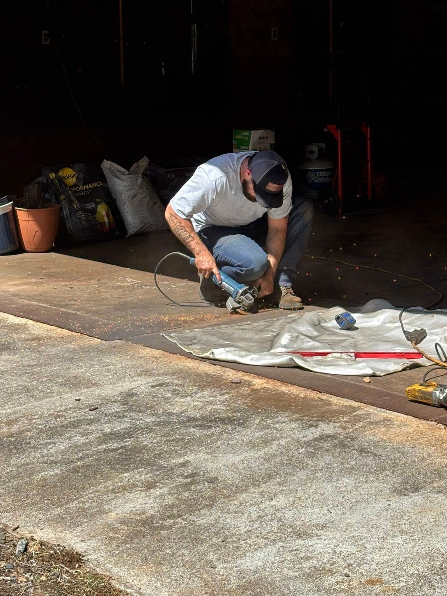 Man in baseball cap and mask using a grinder on the ground, inside a dark garage.