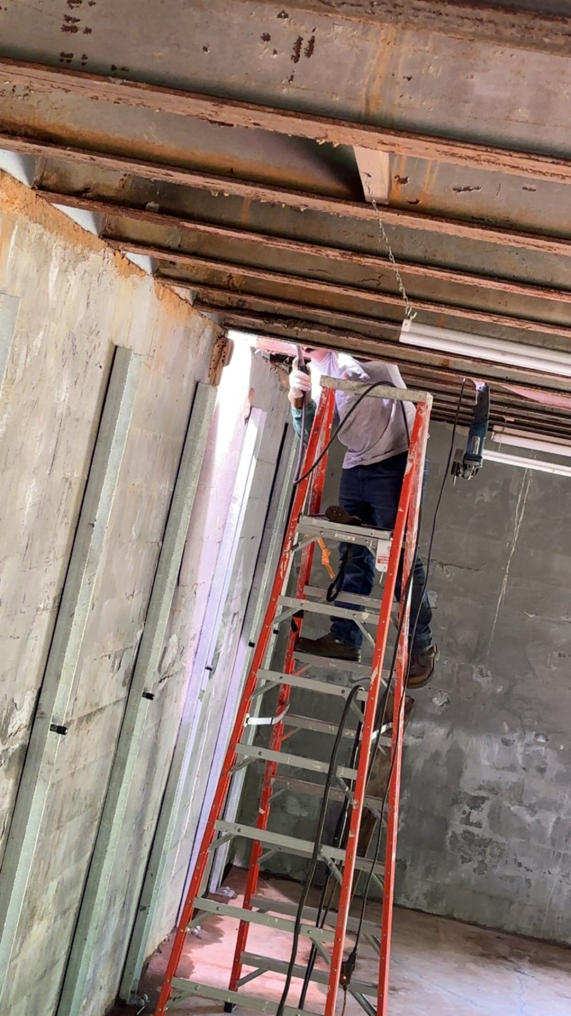 Person on a ladder cutting a material, likely drywall, near a concrete wall and ceiling with exposed beams.