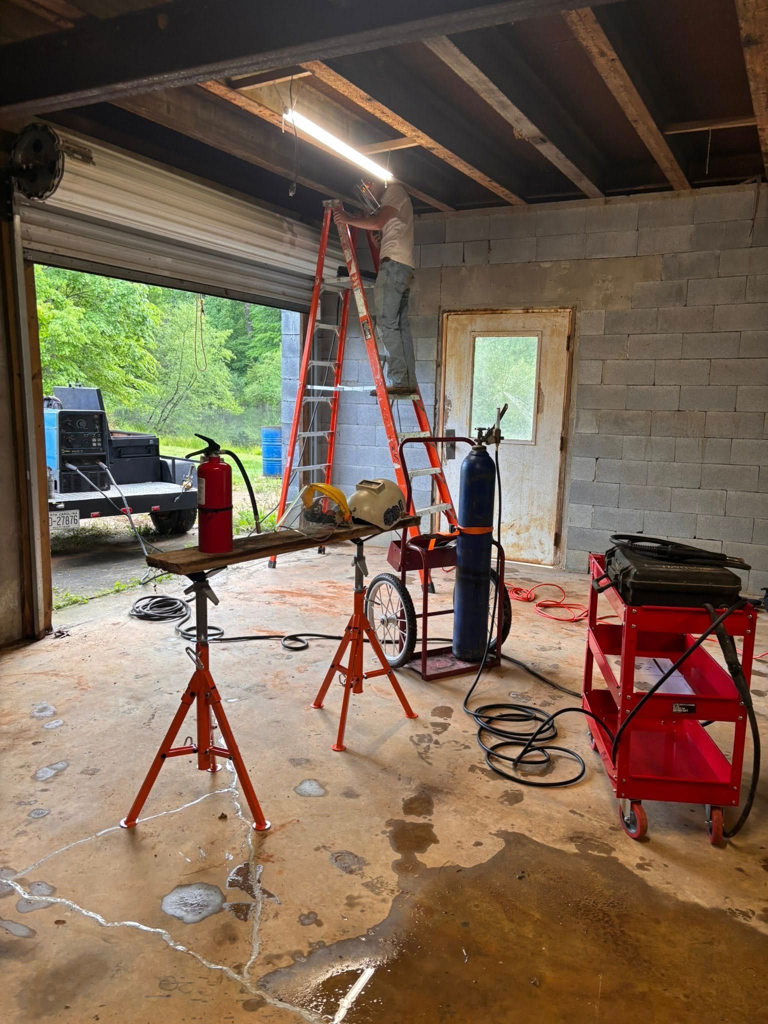Person on ladder, working in a garage. Tools include welding equipment, fire extinguisher, and a sawhorse.