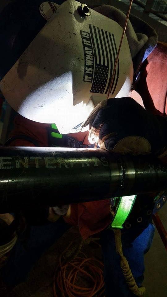 Welder wearing a mask with an American flag, welding a black pipe. Bright sparks are visible.