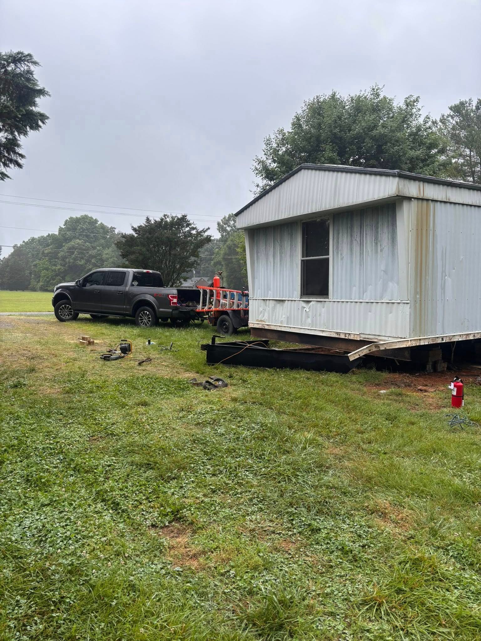 A pickup truck towing a trailer next to a weathered mobile home on a grassy lot. Overcast sky.