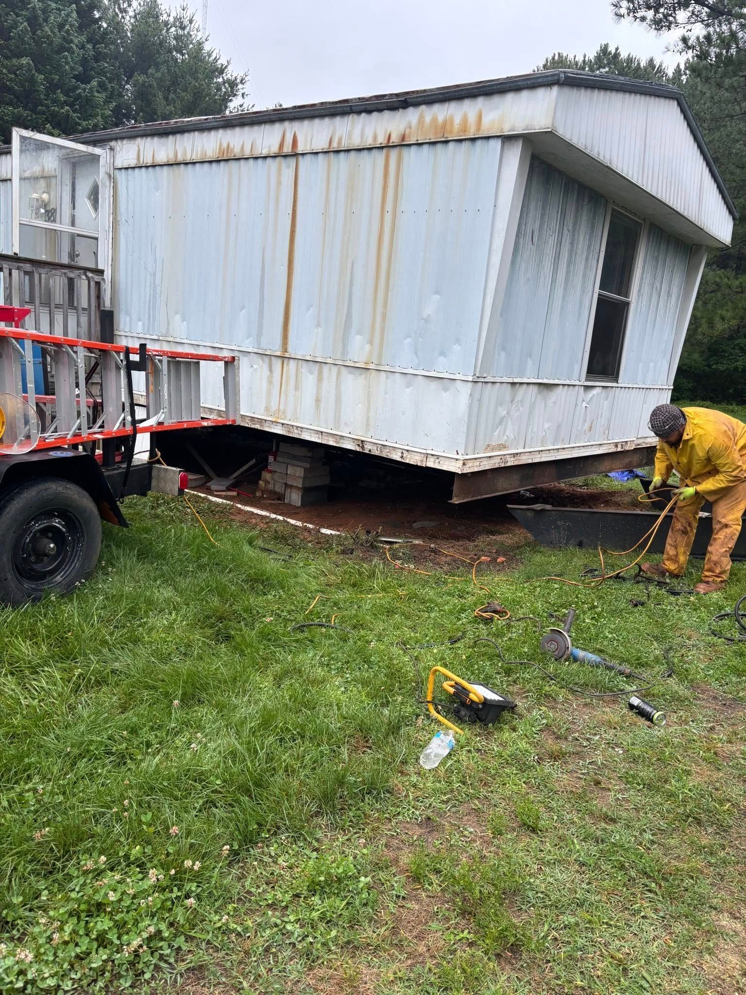 Mobile home tilted, being worked on by a person in protective gear; trailer attached.