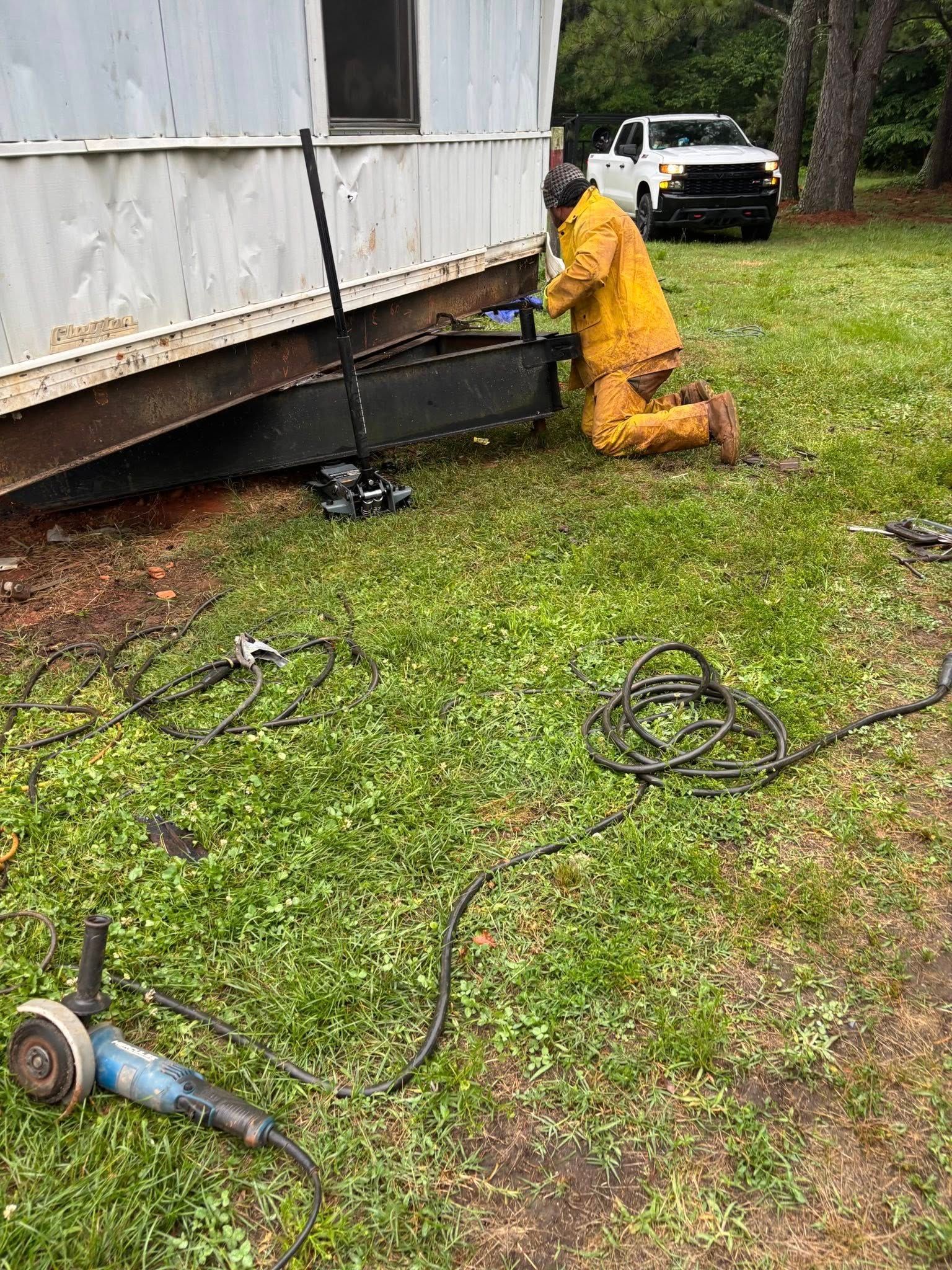 A person in yellow protective gear works on a trailer's metal undercarriage outdoors, next to a white pickup truck.