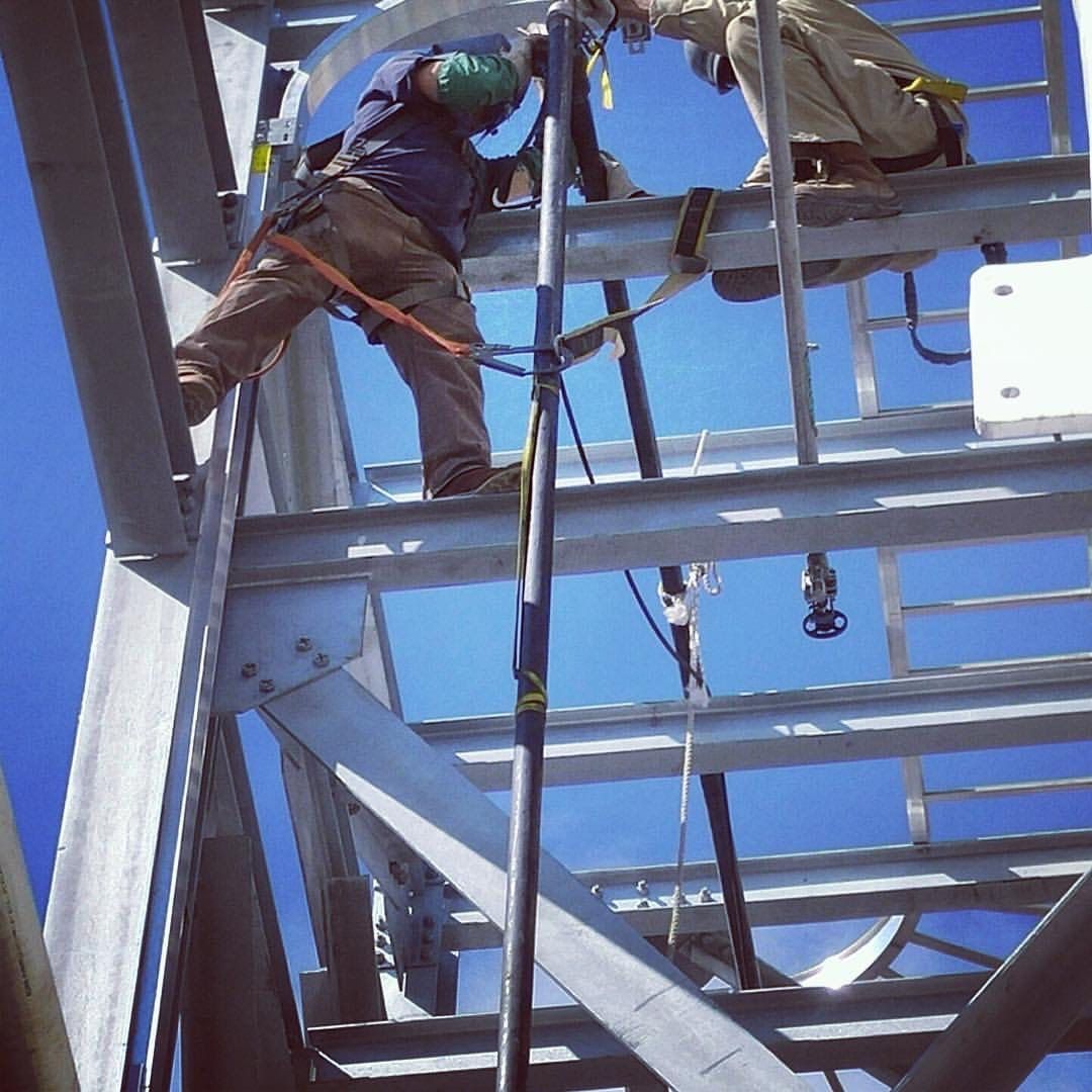 Two workers on metal structure, blue sky, safety harnesses, one attaching a black pole.