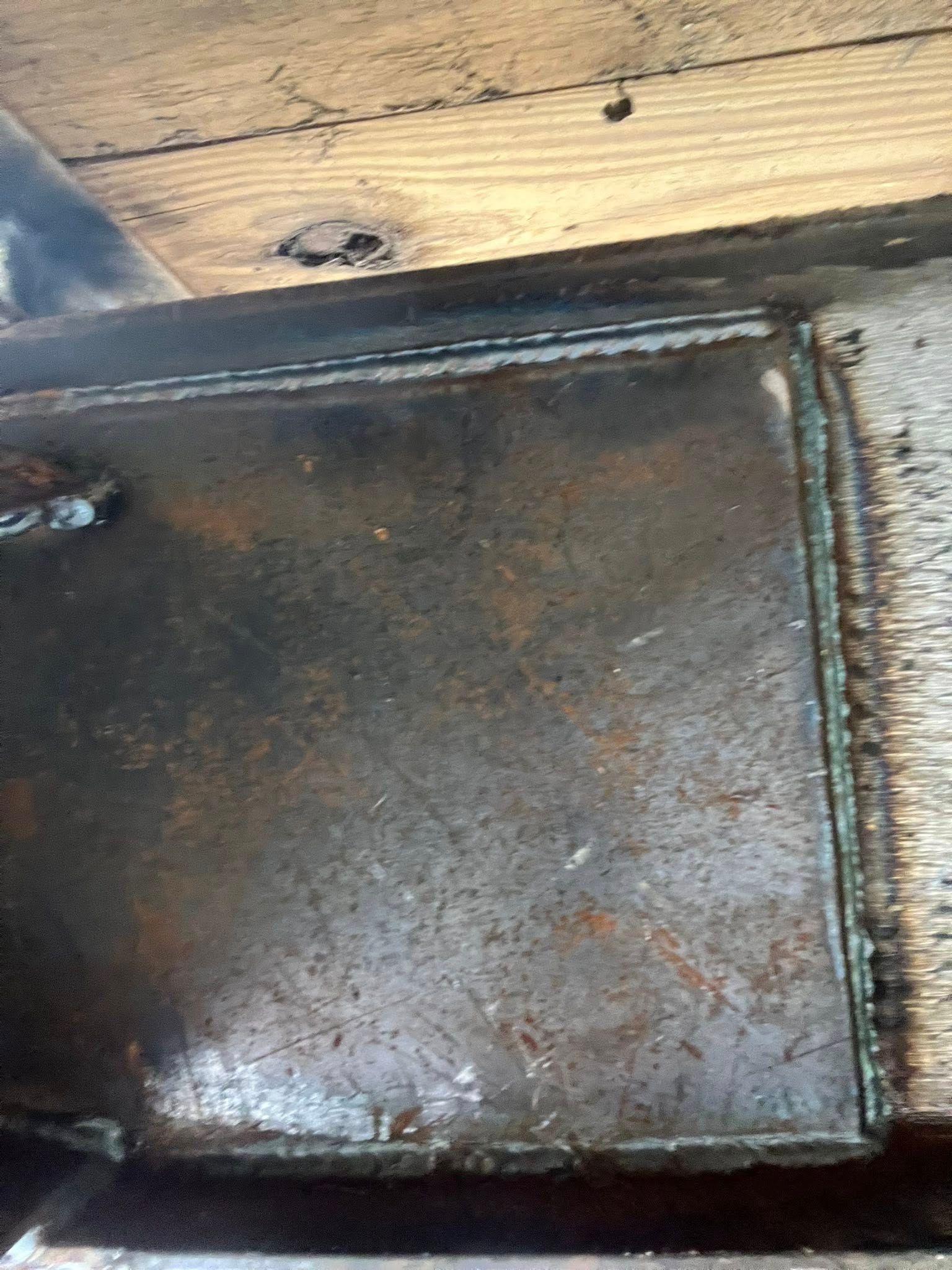 Close-up of a rusted, dark metal plate with square edges, attached to a wooden beam.