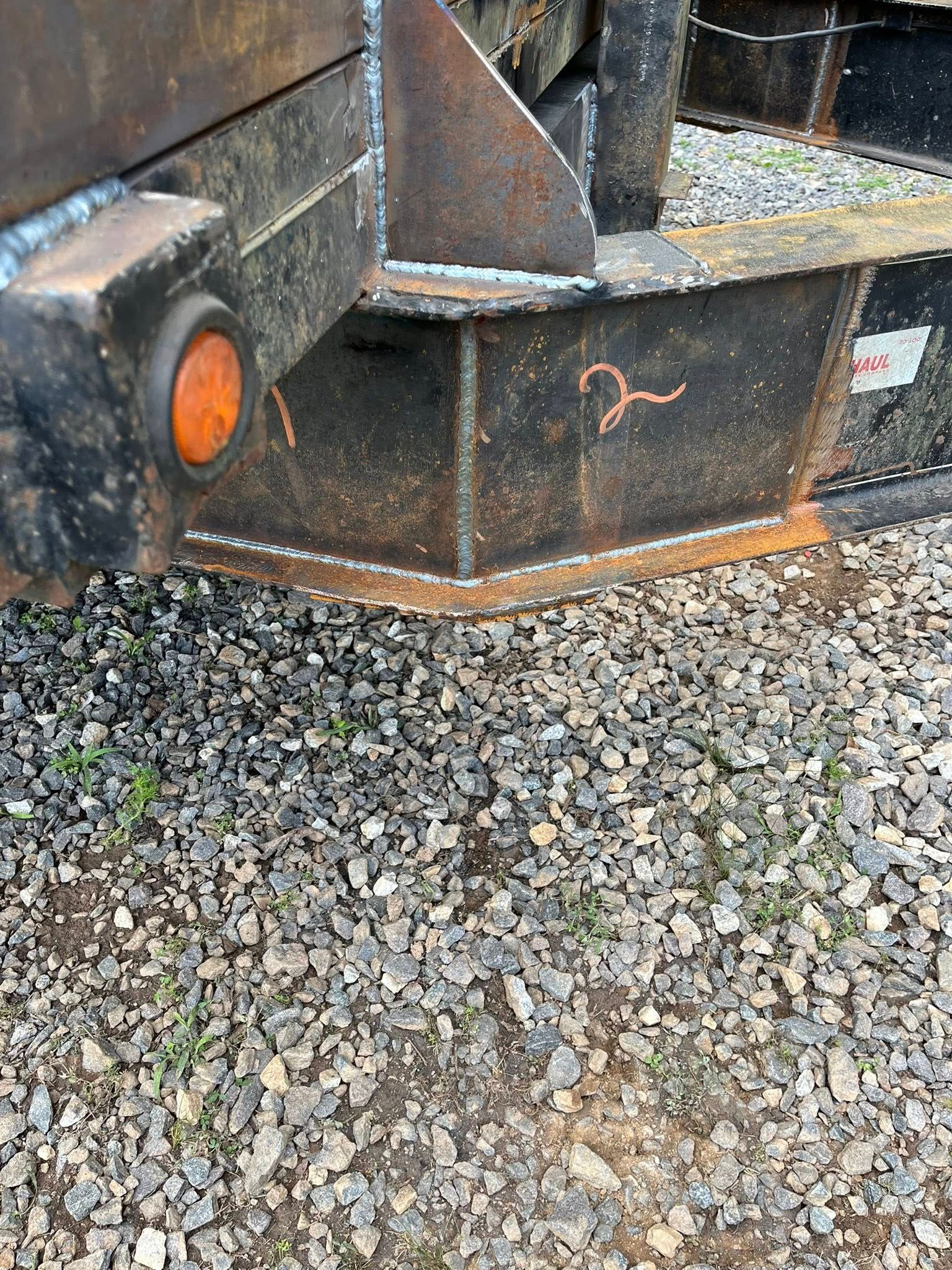 Close-up of a rusty, dark-colored truck bed corner with a round orange light. It is on gravel.