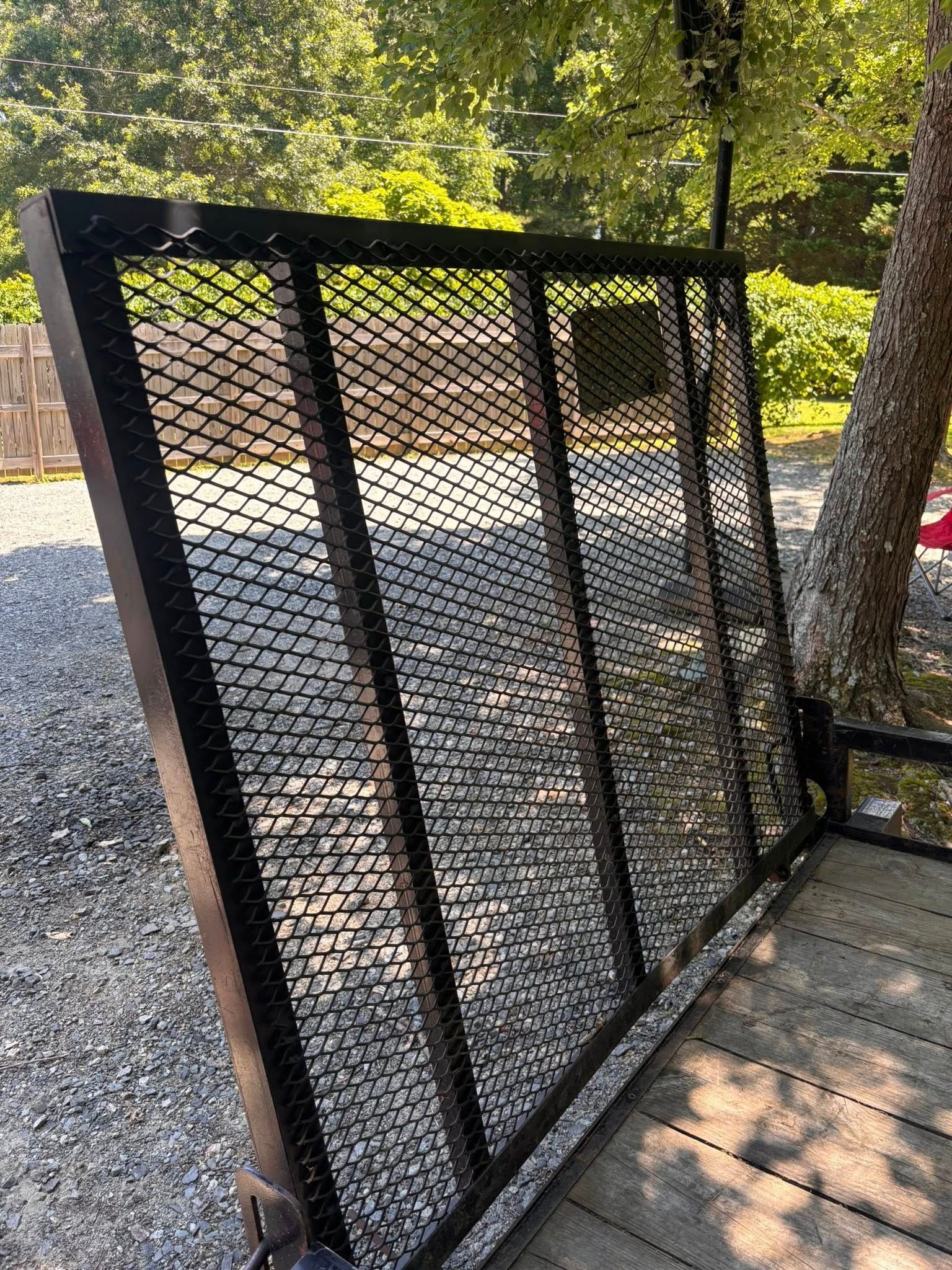 Black metal mesh ramp on a trailer, angled on a gravel surface. Trees and sunlight in the background.