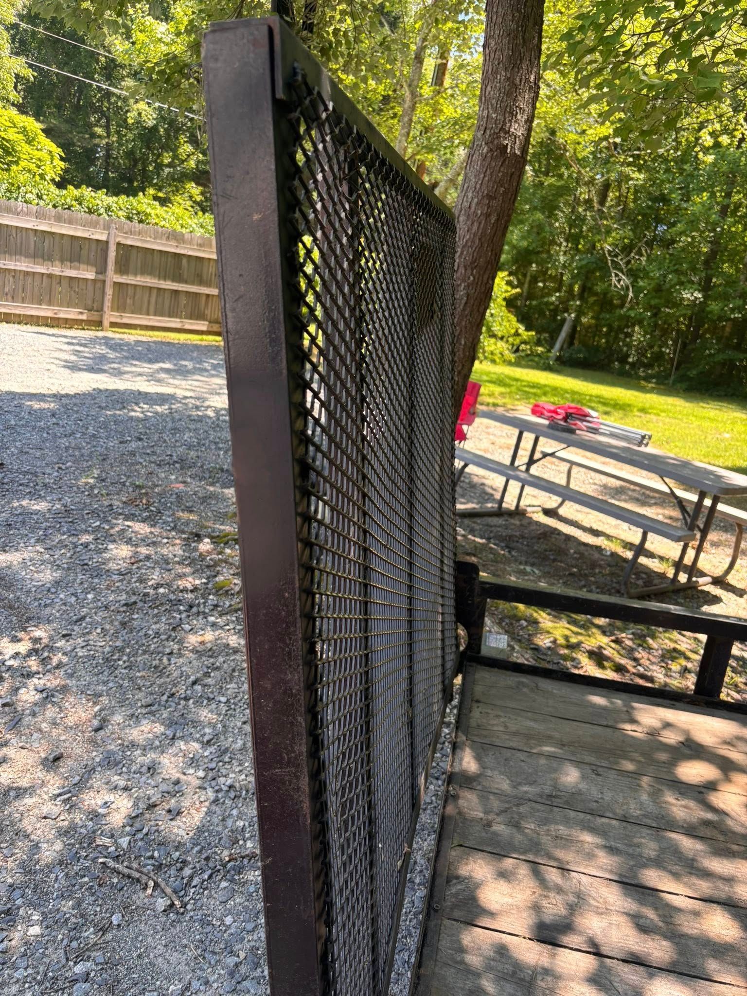 Brown metal mesh trailer side against a gravel driveway with a fence, tree, and picnic table in the background.