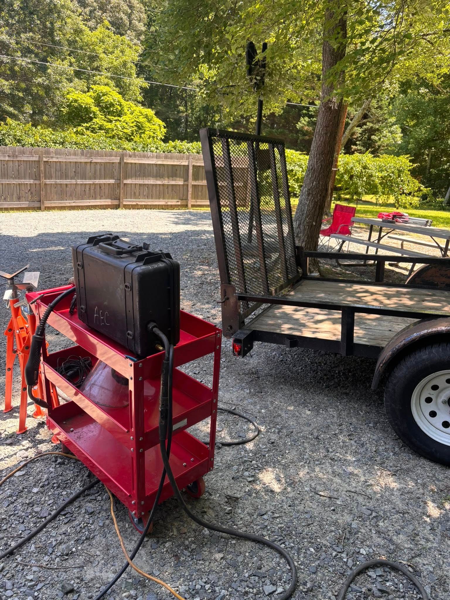Red cart with a black box, cables, and a trailer in a gravel area.