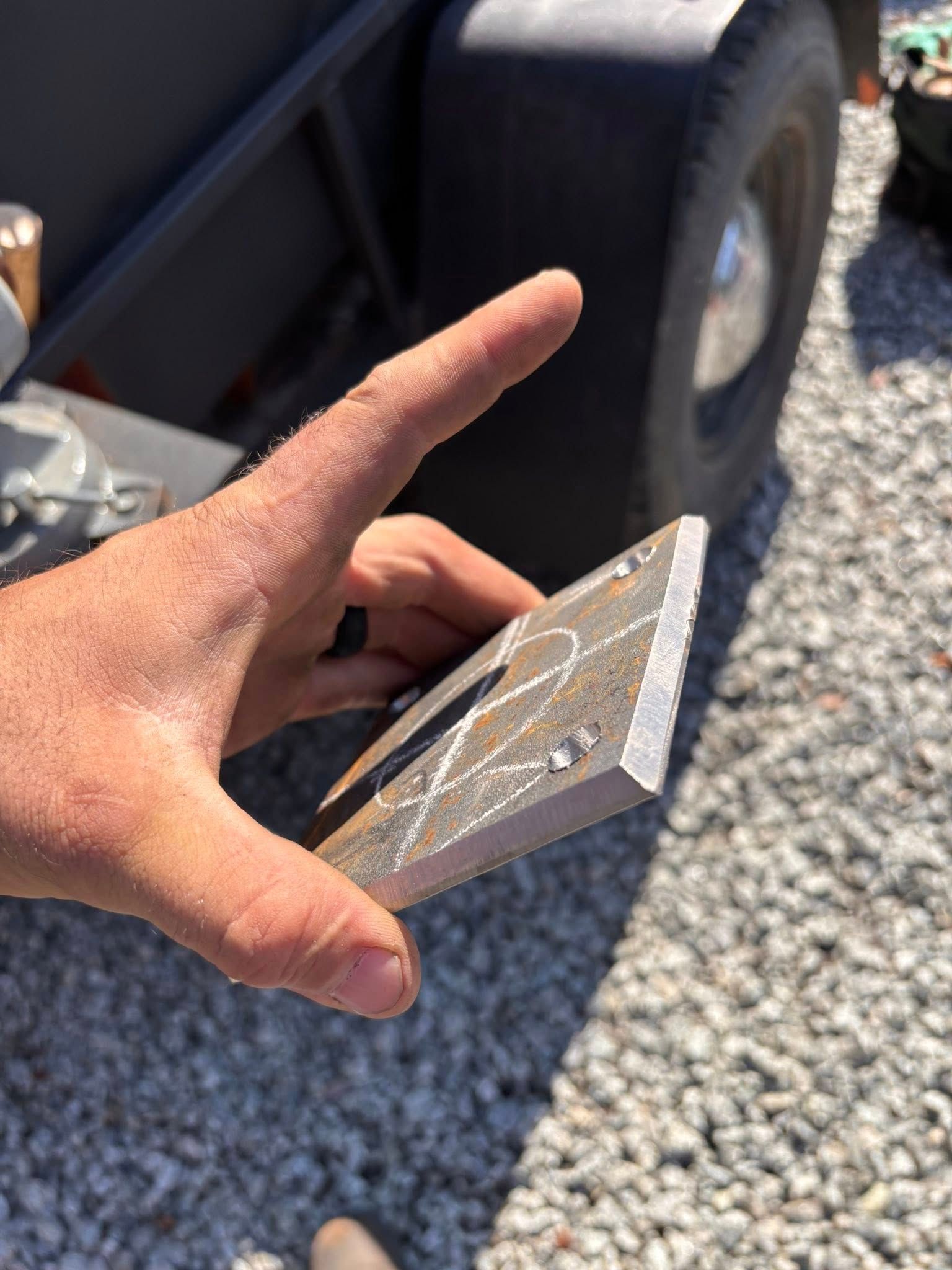 Hand holding a square metal plate with markings, trailer and gravel in background.