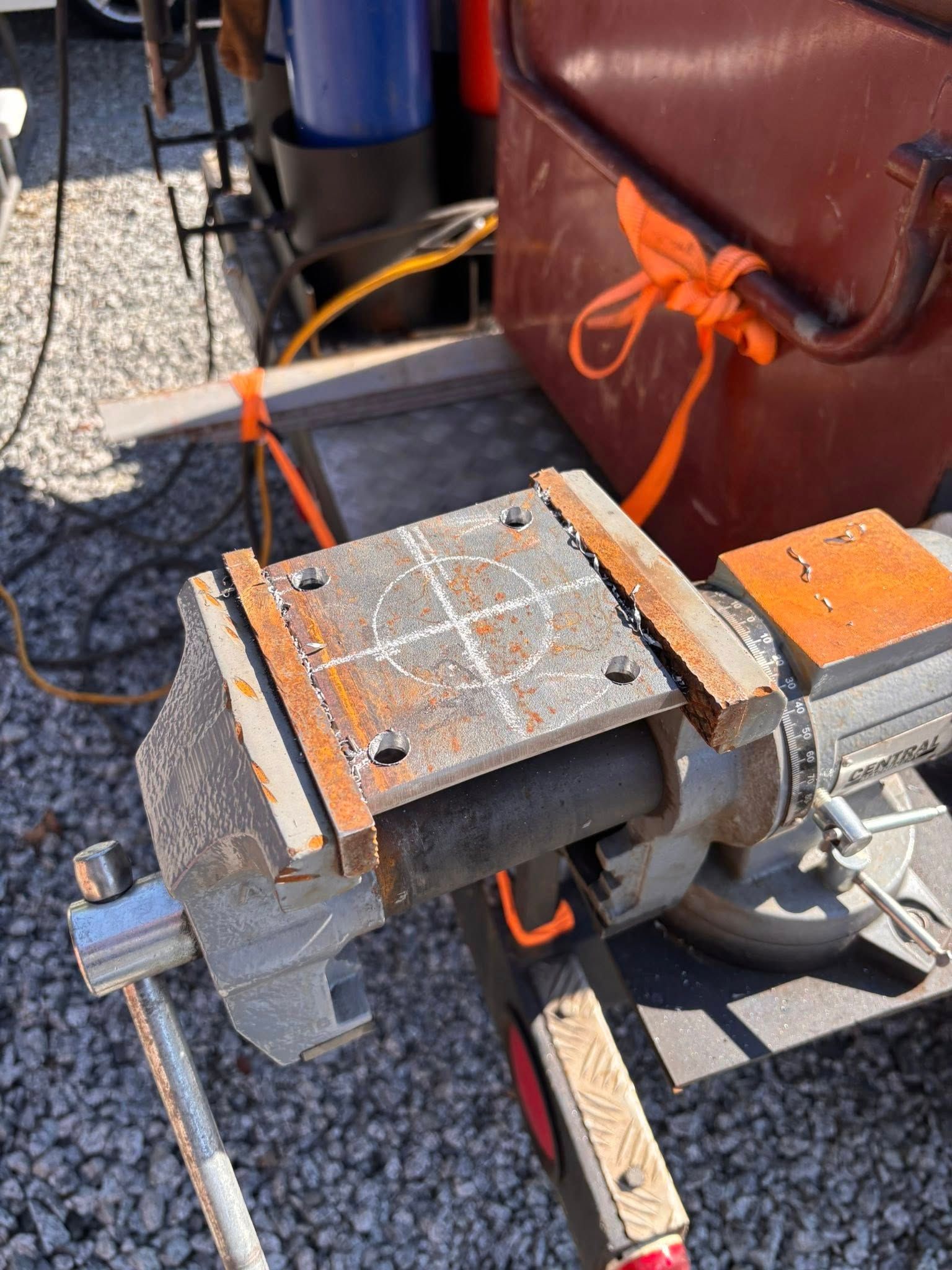 A metal plate clamped in a vise, with lines and markings; brown box and blue tank in background.