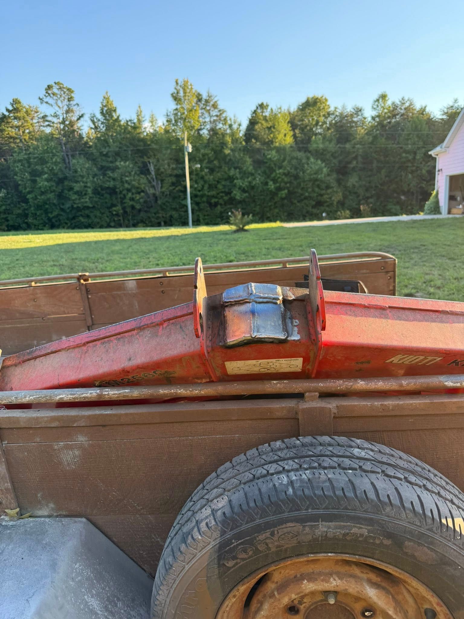 Red and metal machinery mounted on a rusty trailer. A tire is visible on the side, grass and trees in the background.
