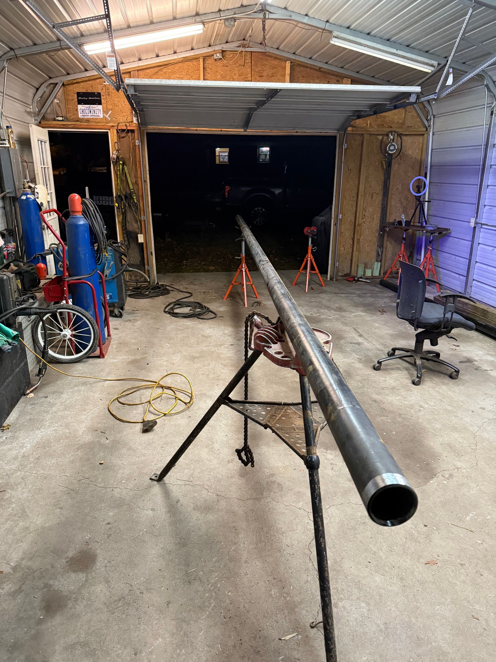A long metal pipe on a stand in a garage workshop. Welding equipment and open garage door in the background.