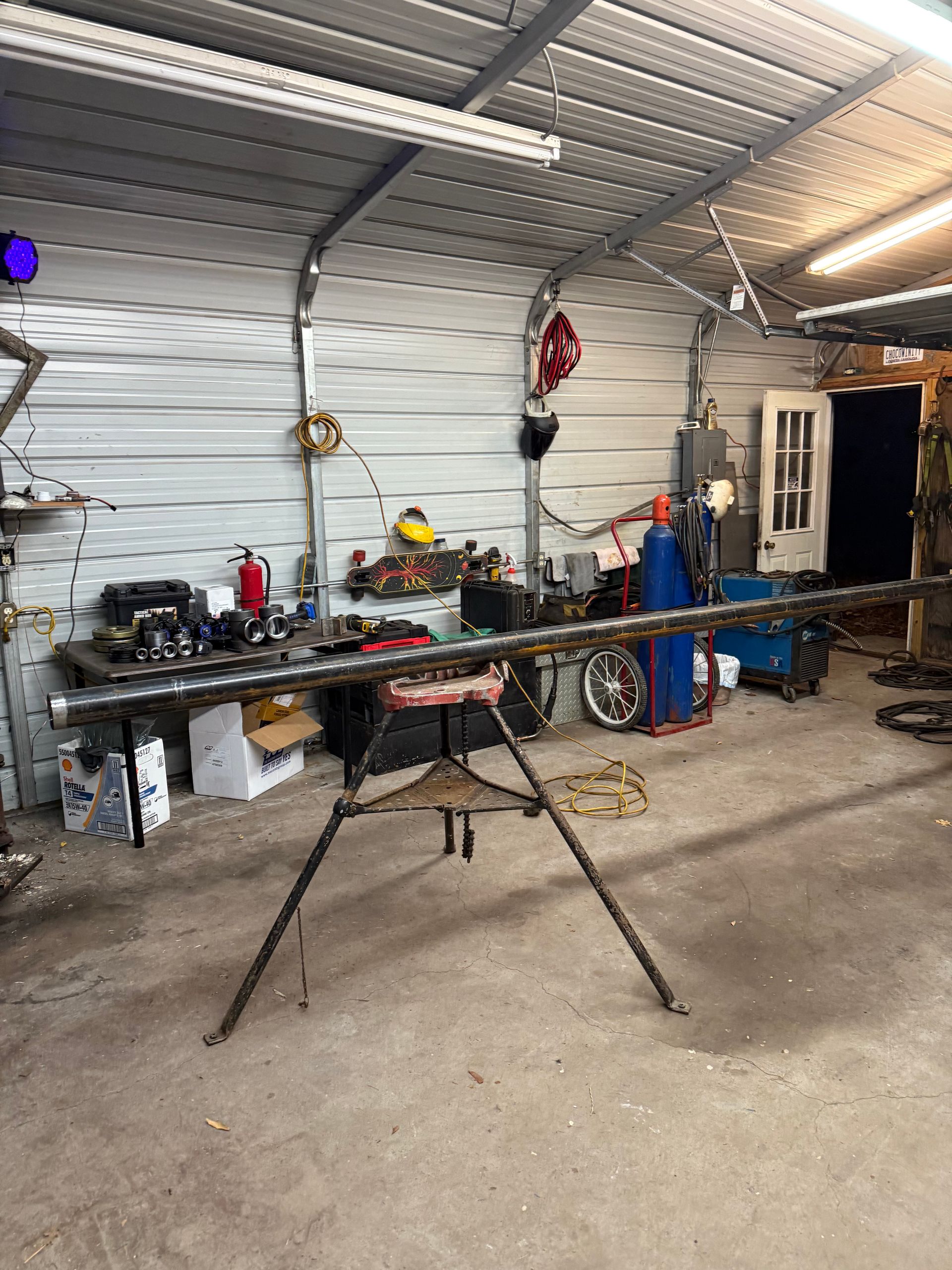 A metal workshop with a long pipe on a tripod stand; tools and equipment scattered throughout.