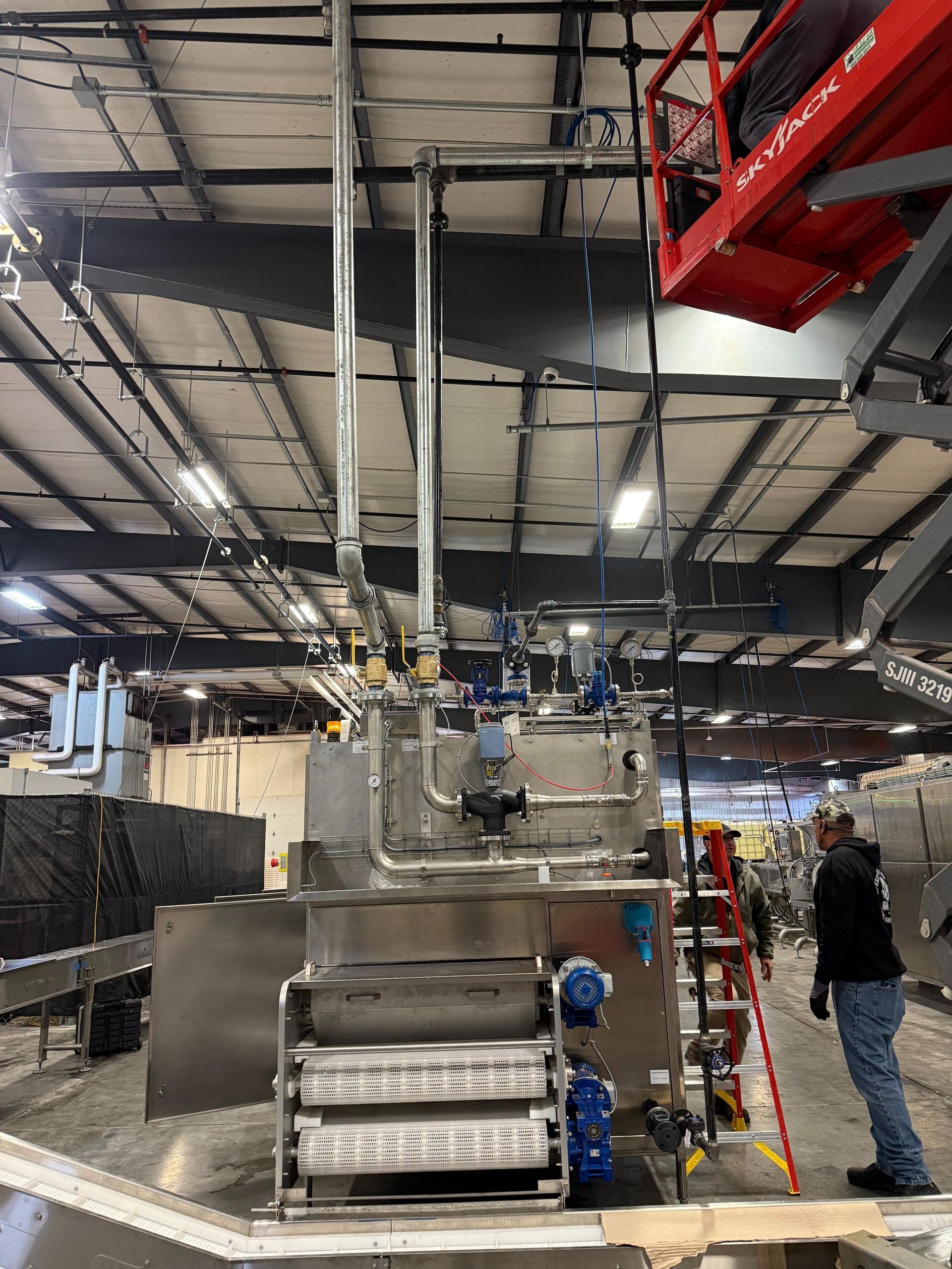 Industrial stainless steel machinery with pipes and valves, indoor setting, a worker, and a lift.