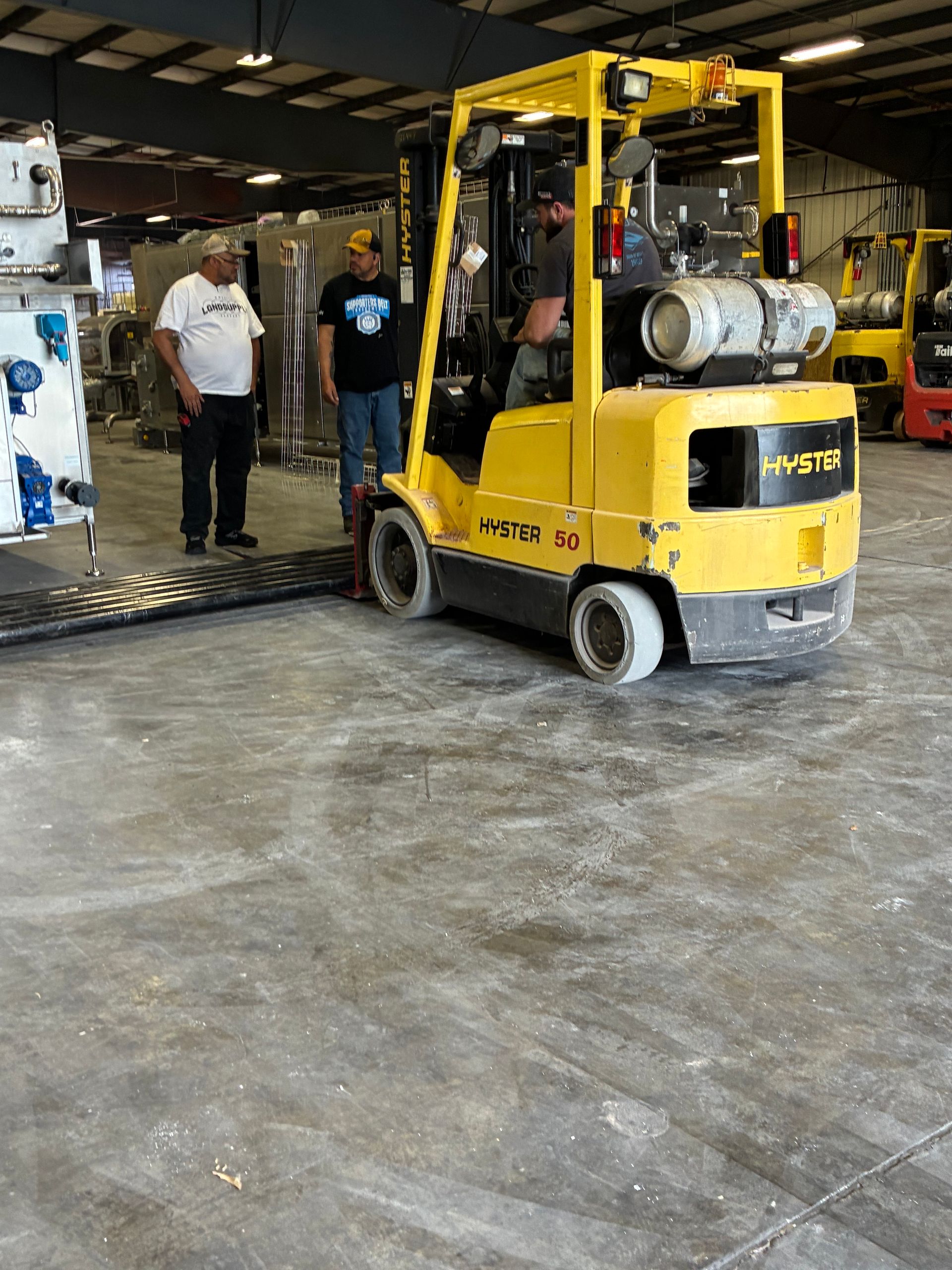 Yellow forklift indoors, operator, two men nearby. Industrial setting, concrete floor.