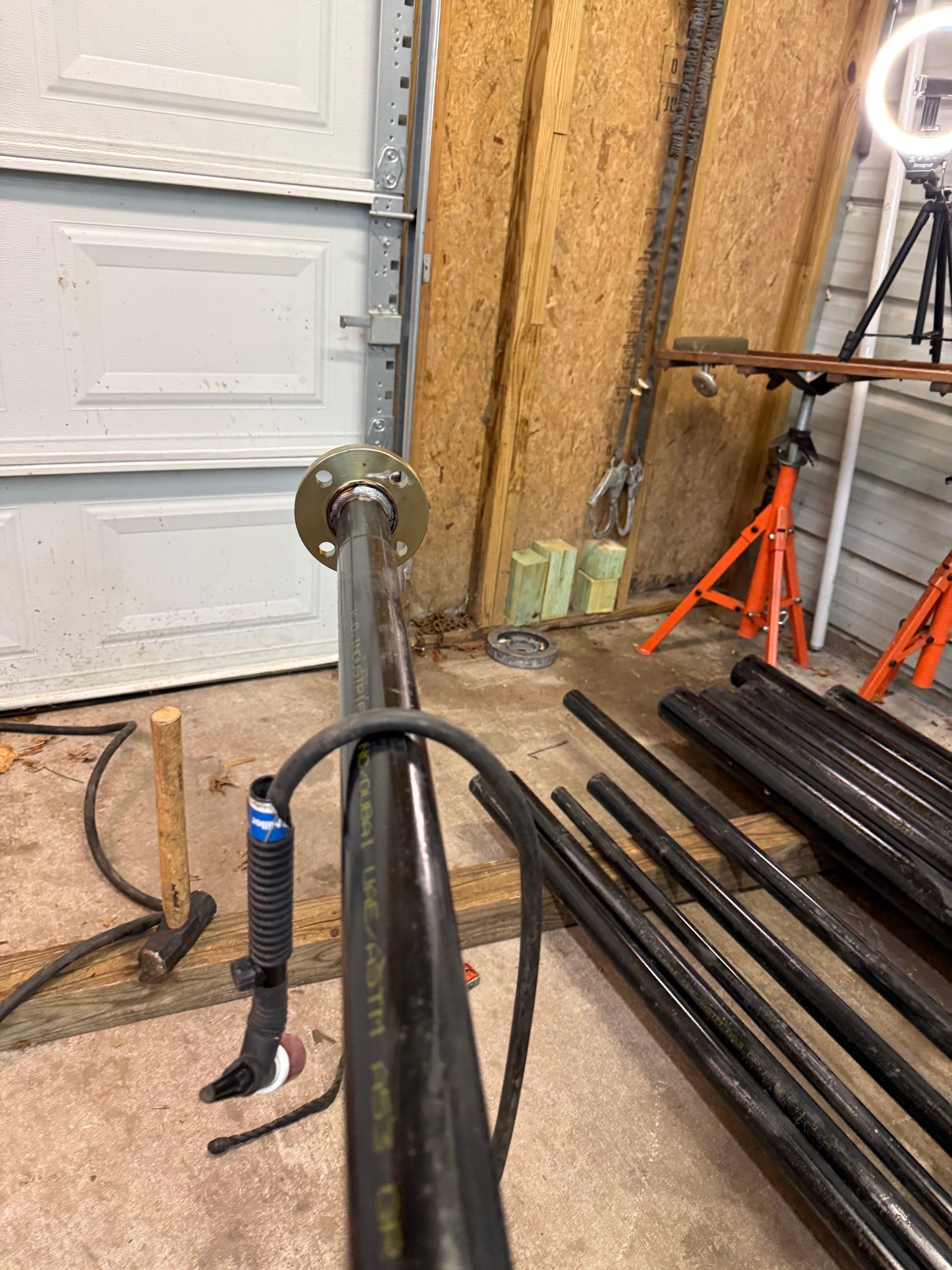 Black metal pipe with a flange, various other pipes, and a welding torch in a garage setting.