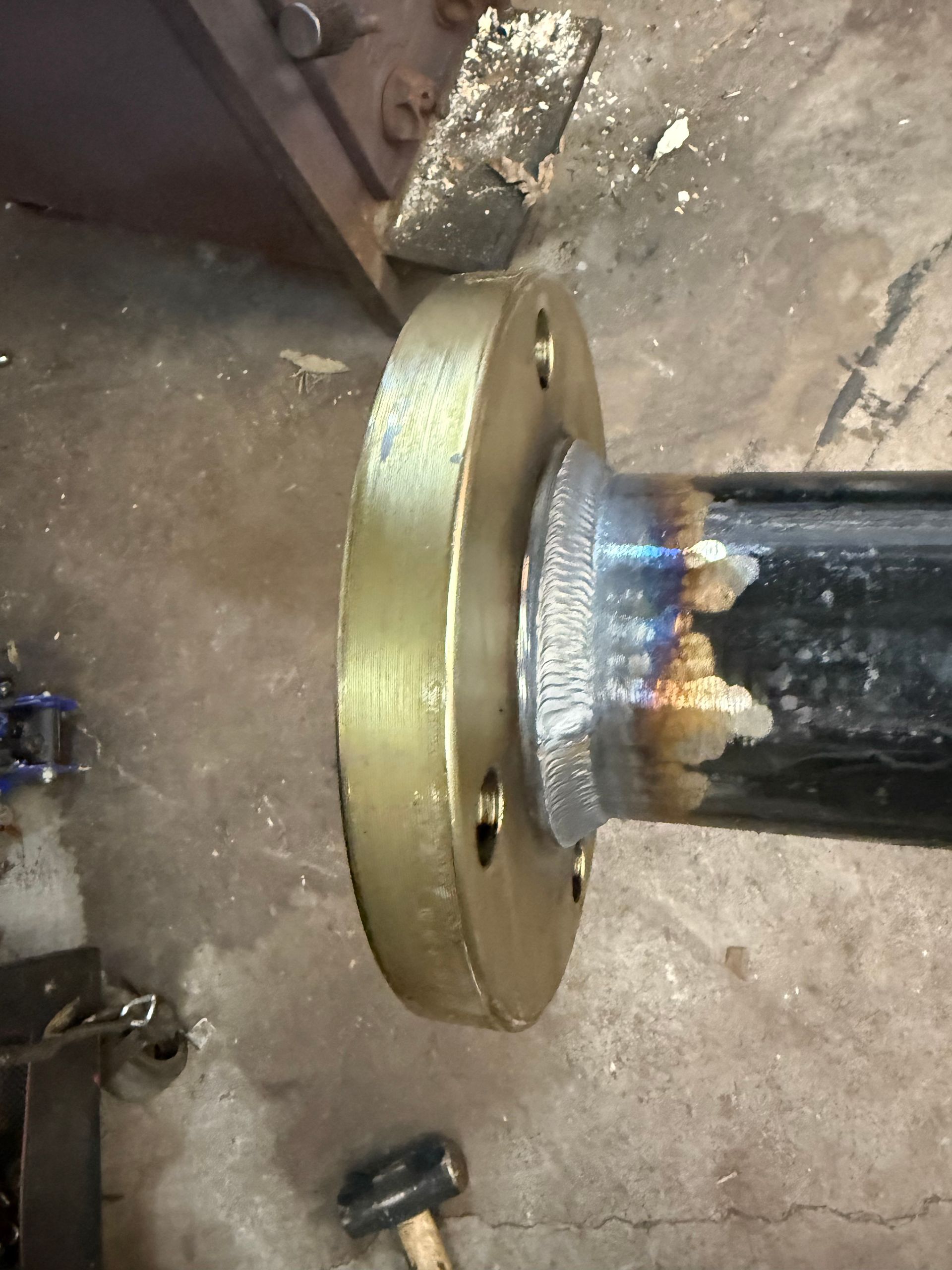 A welded metal flange attached to a black pipe. The flange is gold, with weld discoloration.