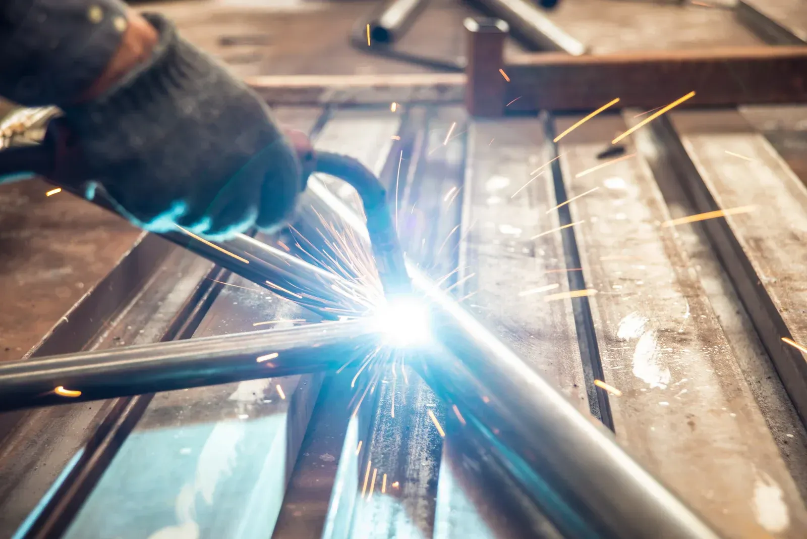 Welder wearing gloves using a welding torch on metal pipes. Sparks and bright light are present.