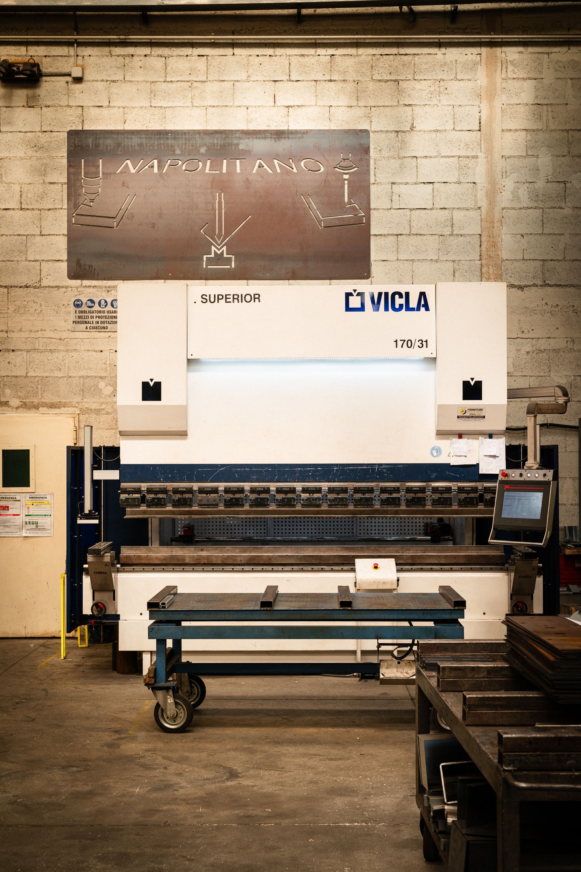 Grande piegatrice per metalli in un'officina. Un carrello blu con lamiere davanti. Sullo sfondo un muro di mattoni.