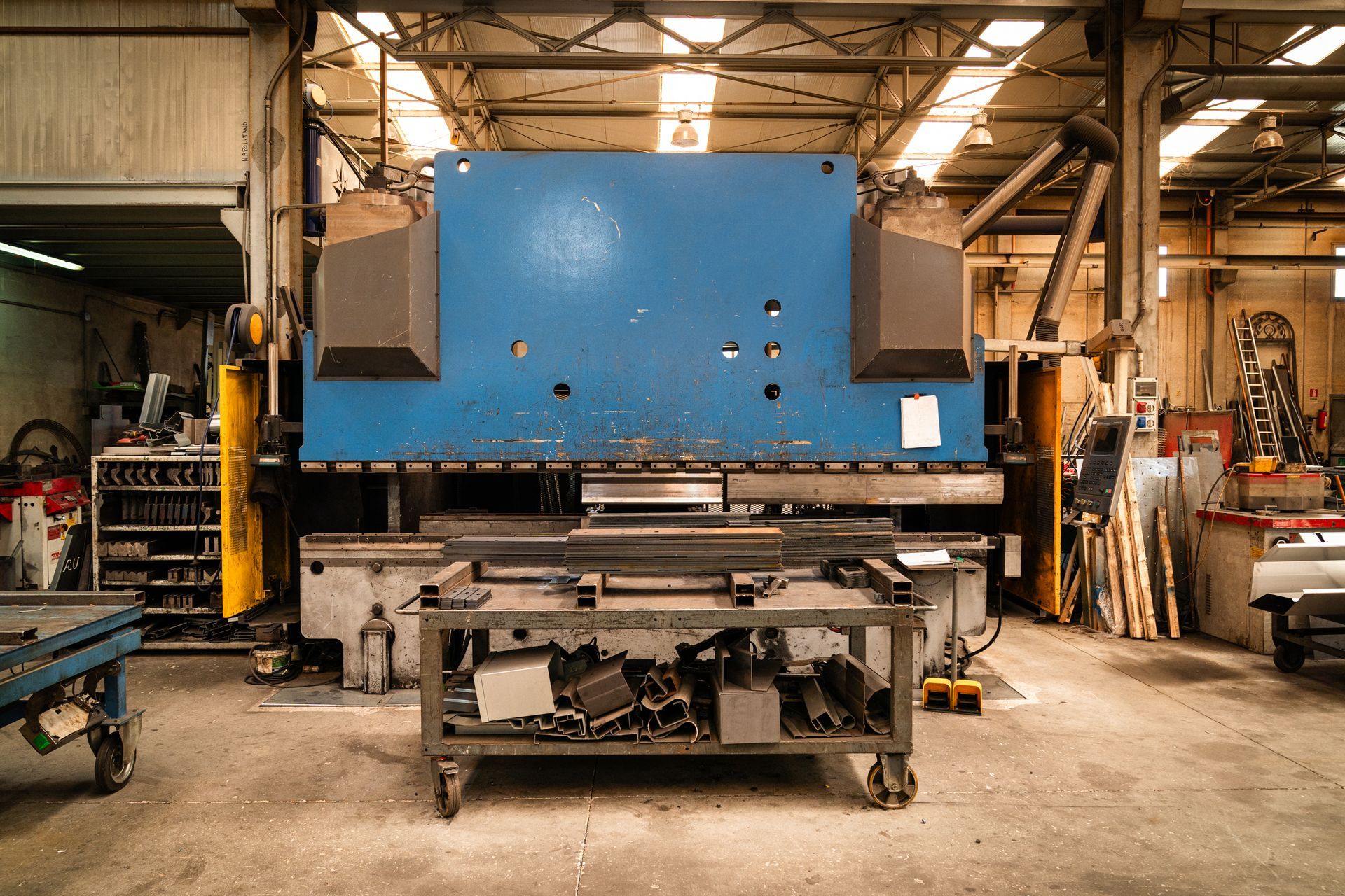 Grande pressa industriale blu in un'officina, con ripiani metallici e utensili visibili.